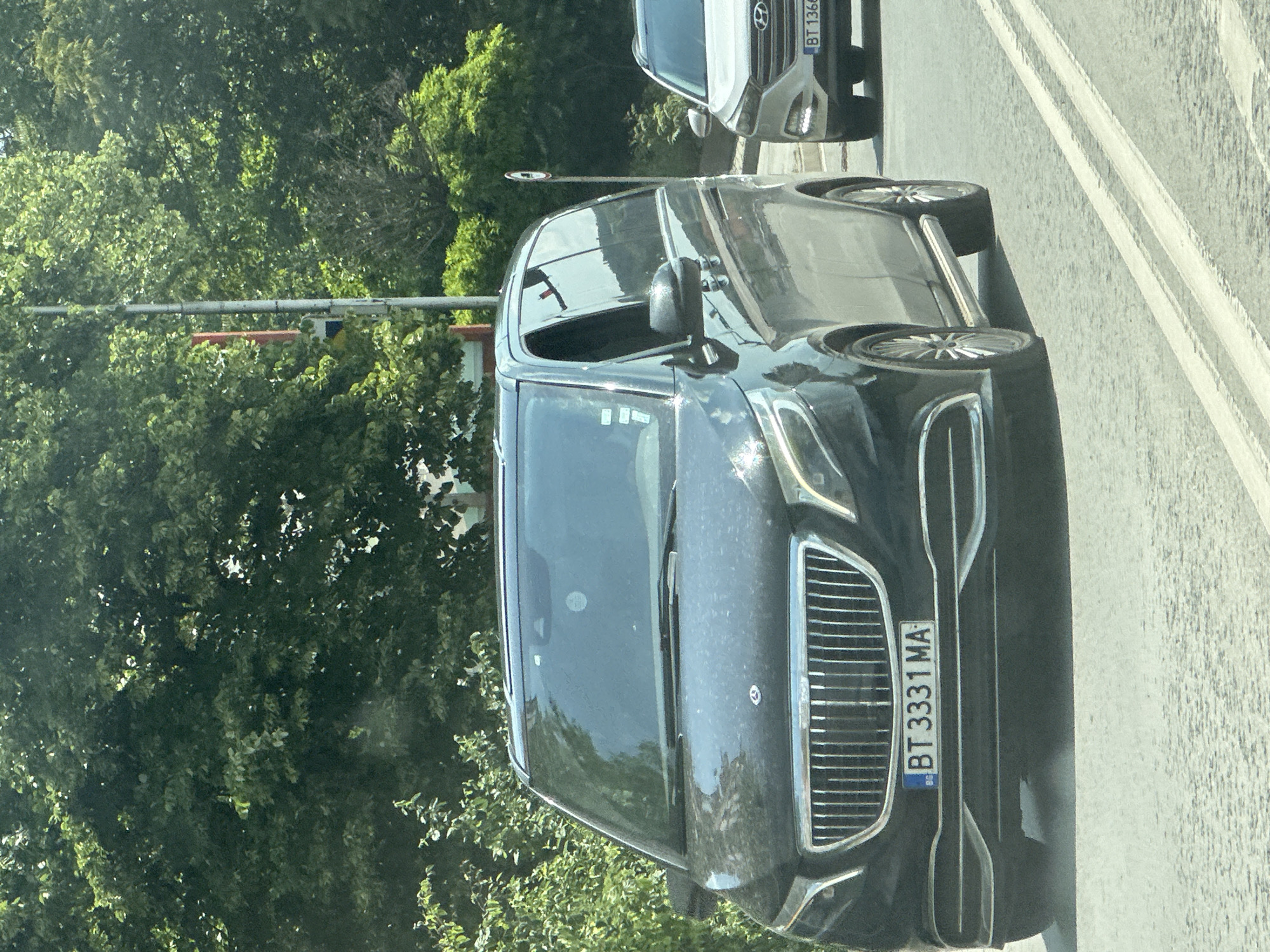The image, taken in Veliko Tarnovo, Bulgaria, captures two vehicles on a sunny day, likely in traffic or parked on a street lined with lush green trees. The photograph is oriented vertically, showing the vehicles from a slightly elevated perspective.
In the foreground, a dark grey or black minivan or large SUV is prominently featured, facing the viewer. It displays a large, vertical chrome grille and distinctive C-shaped daytime running lights. A small, stylized emblem is visible on the hood, possibly an 'M' or a specific brand logo. The vehicle's windshield reflects the bright sky and parts of the surrounding environment, including a circular "No Entry" or "No Stopping" sign. The car's front license plate clearly reads "BG BT 3331 MA", indicating it is registered in Bulgaria, specifically the Veliko Tarnovo district (BT). The vehicle is equipped with multi-spoke alloy wheels.
Behind the foreground car and to its right, a lighter-colored SUV, possibly silver or white, is partially visible. Its front bumper and a portion of its grille are seen, along with its Bulgarian license plate, which reads "BT 1360". The "BT" prefix further confirms the Veliko Tarnovo location.
The background is dominated by dense, vibrant green foliage from numerous trees, suggesting a pleasant, natural environment amidst the road. The road surface itself is visible to the right of the cars, featuring white painted lines. The bright sunlight indicates it is daytime, and the overall impression is one of clear and fair weather. No people are visible in the scene.