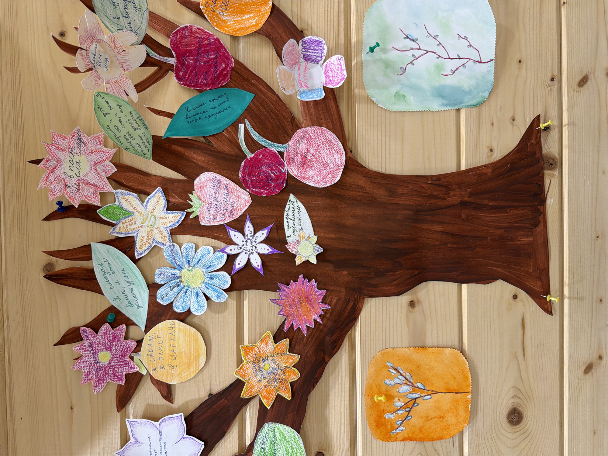 The image shows a bulletin board made of light-colored wooden planks. Affixed to the board is a hand-painted, brown tree with various paper cutouts attached to its branches. These cutouts include flowers, fruits (cherries and strawberries), leaves, and circles, all colored and decorated with hand-drawn patterns and written text in Russian. A few of the cutouts are attached with colorful push pins. Two additional square paper images are attached to the board near the tree, one depicting a branch with buds and the other a twig with a bird. The overall impression is of a child's art project or a collaborative display.