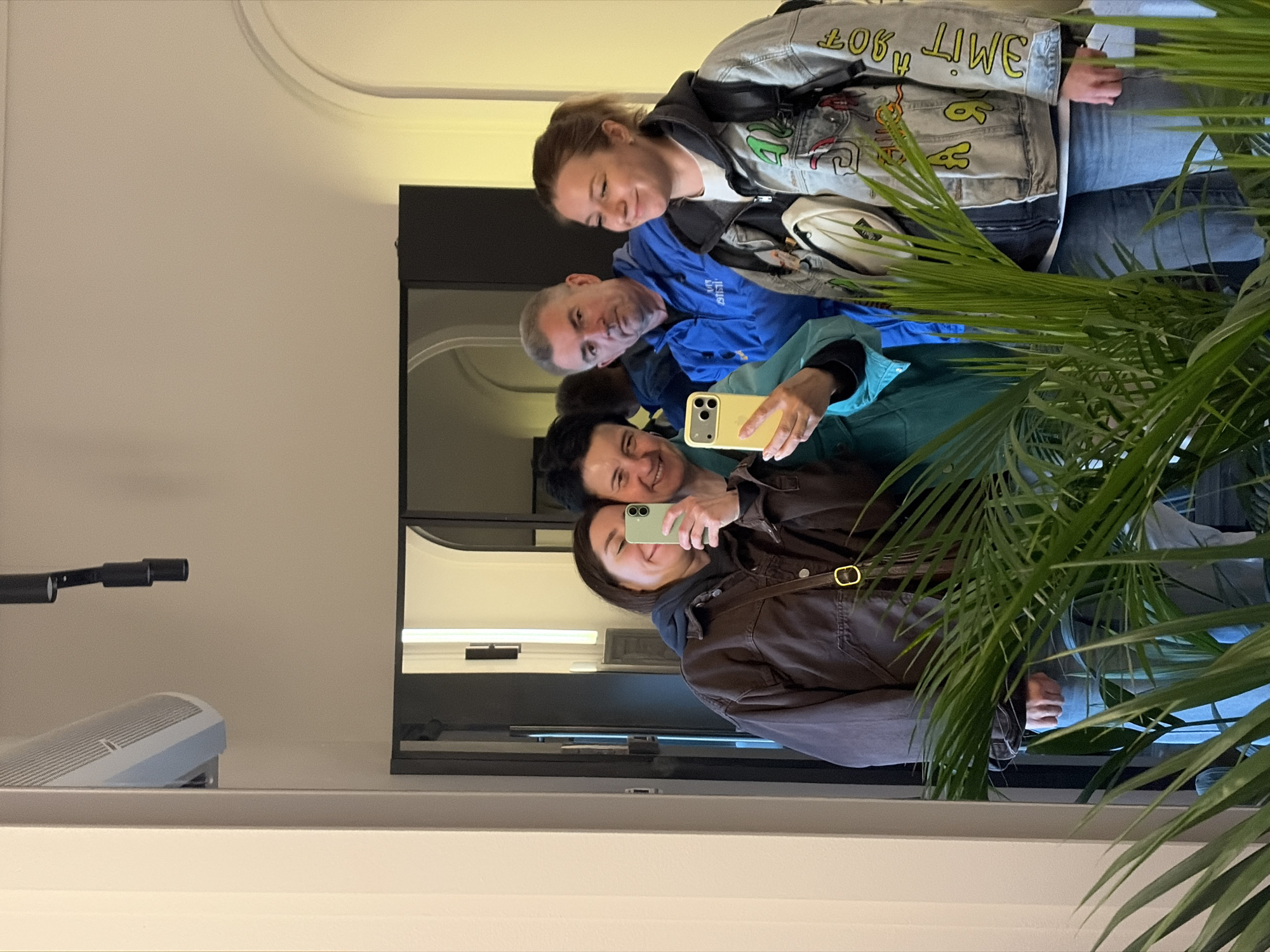 Four individuals are captured in a selfie taken in front of a mirror. The person taking the selfie has a brown jacket on. Two others are directly behind her, and the fourth is partially visible, wearing a denim jacket. They are indoors, as seen from the architecture reflected in the mirror. A plant with large green leaves is partially obscuring the view on the right side of the frame. An air conditioning unit and a microphone are visible near the mirror's edge. The location is most likely Бараўлянскі сельскі Савет, Belarus.