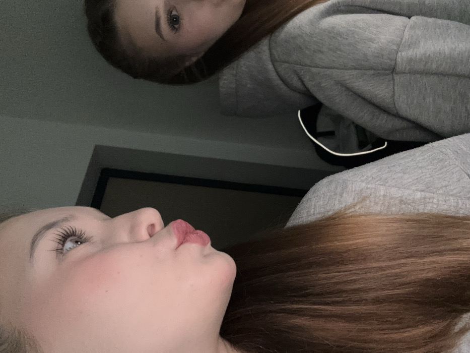 Two women are captured in this selfie-style image. The woman in the foreground faces upwards with pursed lips. She has fair skin, long eyelashes, and auburn hair. Above her, the second woman, wearing a gray hoodie, looks down into the camera. The background reveals a dark door frame and a dimly lit room. The image has a casual, candid feel.