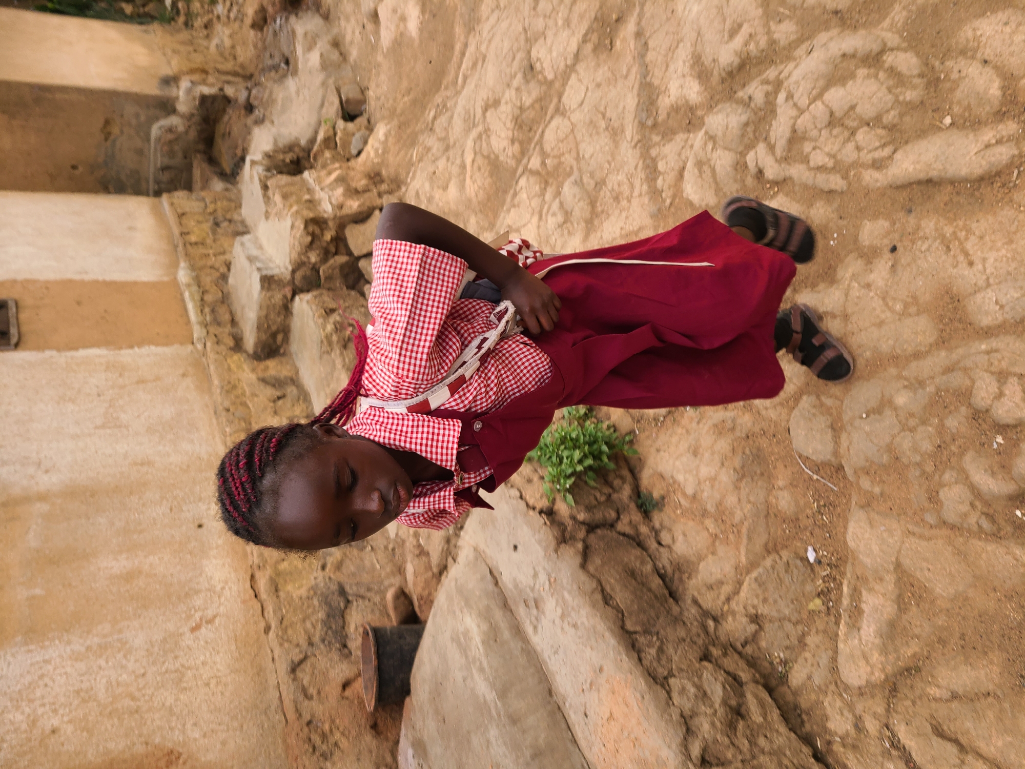A young girl is shown standing outdoors, viewed from a slightly elevated perspective. She has dark skin and her hair is braided with a mix of red and dark strands. She is wearing a long-sleeved red and white checkered shirt, possibly a school uniform, over a plain dark red skirt. On her feet are dark, strapped sandals. Her eyes are closed or cast downwards, giving her a calm and contemplative appearance.
The setting appears to be a rustic outdoor area with uneven, light brown ground composed of earth, sand, and embedded rocks, showing natural cracks and textures. In the background to the left, there are light-colored, rough concrete or stucco structures, possibly walls or steps of a building. A dark, cylindrical object, resembling a pipe, is partially visible near the bottom left. Small clusters of green foliage sprout from the ground in various spots.
The lighting suggests it is daytime, with soft, diffused illumination that avoids harsh shadows, possibly indicating an overcast sky or an early/late hour. The scene depicts a quiet moment in what appears to be a local, unpaved environment. No visible text is present in the image. The location context provided is Jos, Nigeria.