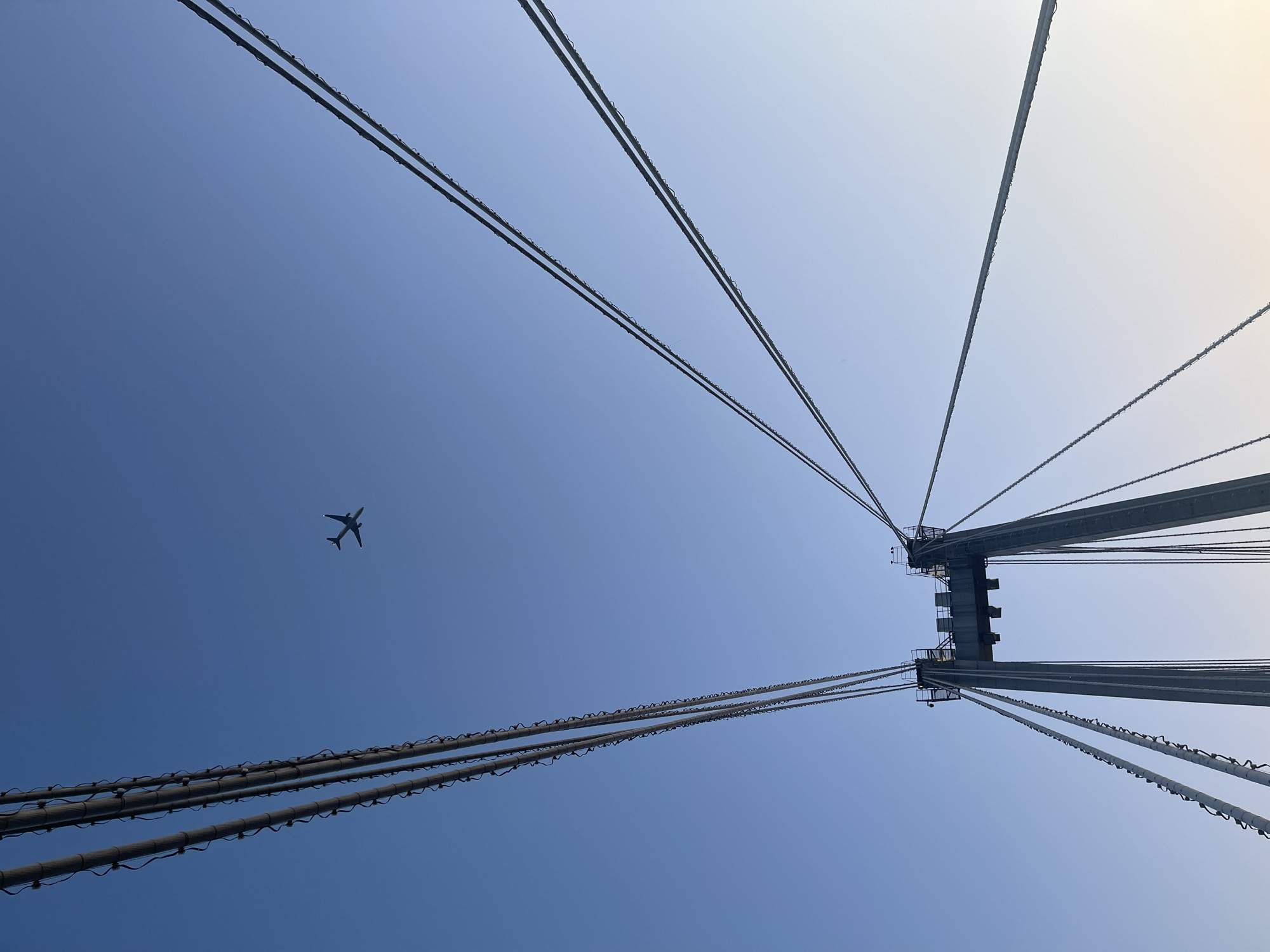 The image shows a low-angle view of a bridge structure with suspension cables against a clear blue sky. An airplane is visible in the upper portion of the frame, flying to the left. The bridge appears to be the Yenisei Bridge in Krasnoyarsk, Russia, known for its distinctive cable-stayed design. The cables are thick and numerous, stretching upwards and outwards from a central tower. The tower itself is a large, grey concrete structure with visible metal components and platforms at the top. The sky transitions from a deep blue at the top of the image to a lighter hue towards the horizon, suggesting it may be early morning or late afternoon. The overall impression is one of scale and engineering, with the airplane adding a sense of depth and movement to the scene.