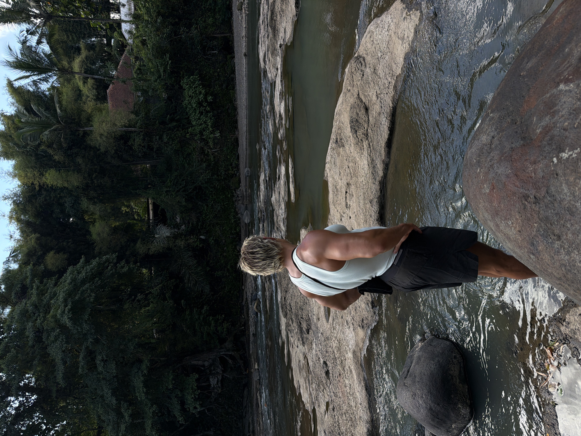 A man stands on rocks in a river in Selemadeg Timur, Indonesia. He wears a white tank top and dark shorts. The river flows over large rocks and through a dense forest.