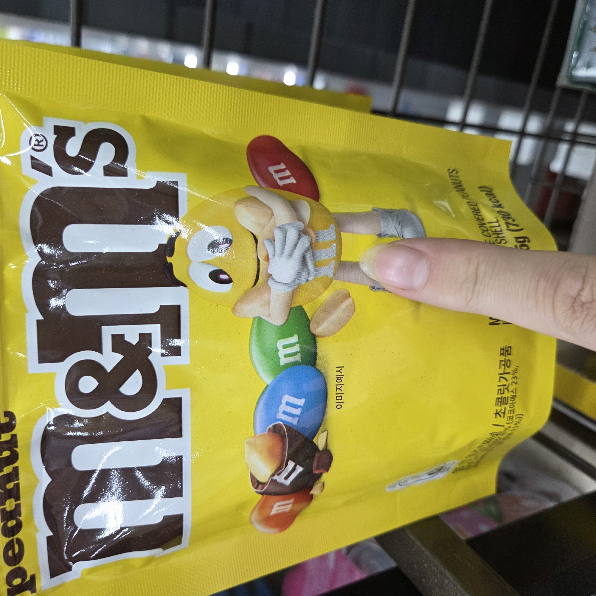 In Dunnae-myeon, South Korea, a yellow M&M's candy bag is displayed. The bag prominently features the M&M's logo in large brown letters on a white background, with a registered trademark symbol above the "s." Several M&M's candies, depicted as animated characters, are shown on the bag. One M&M character, a yellow one with arms crossed, is central. Surrounding it are cartoonish representations of M&M's candies in orange, blue, and green, each with an "m" printed on them. A broken M&M candy is also shown, revealing a peanut inside. Text on the bag is in both English and Korean. A human finger is visible in the foreground, touching the bag. The background appears to be a store shelf with metal grates and out-of-focus items, suggesting the bag is on display for purchase. The lighting is bright, illuminating the product.