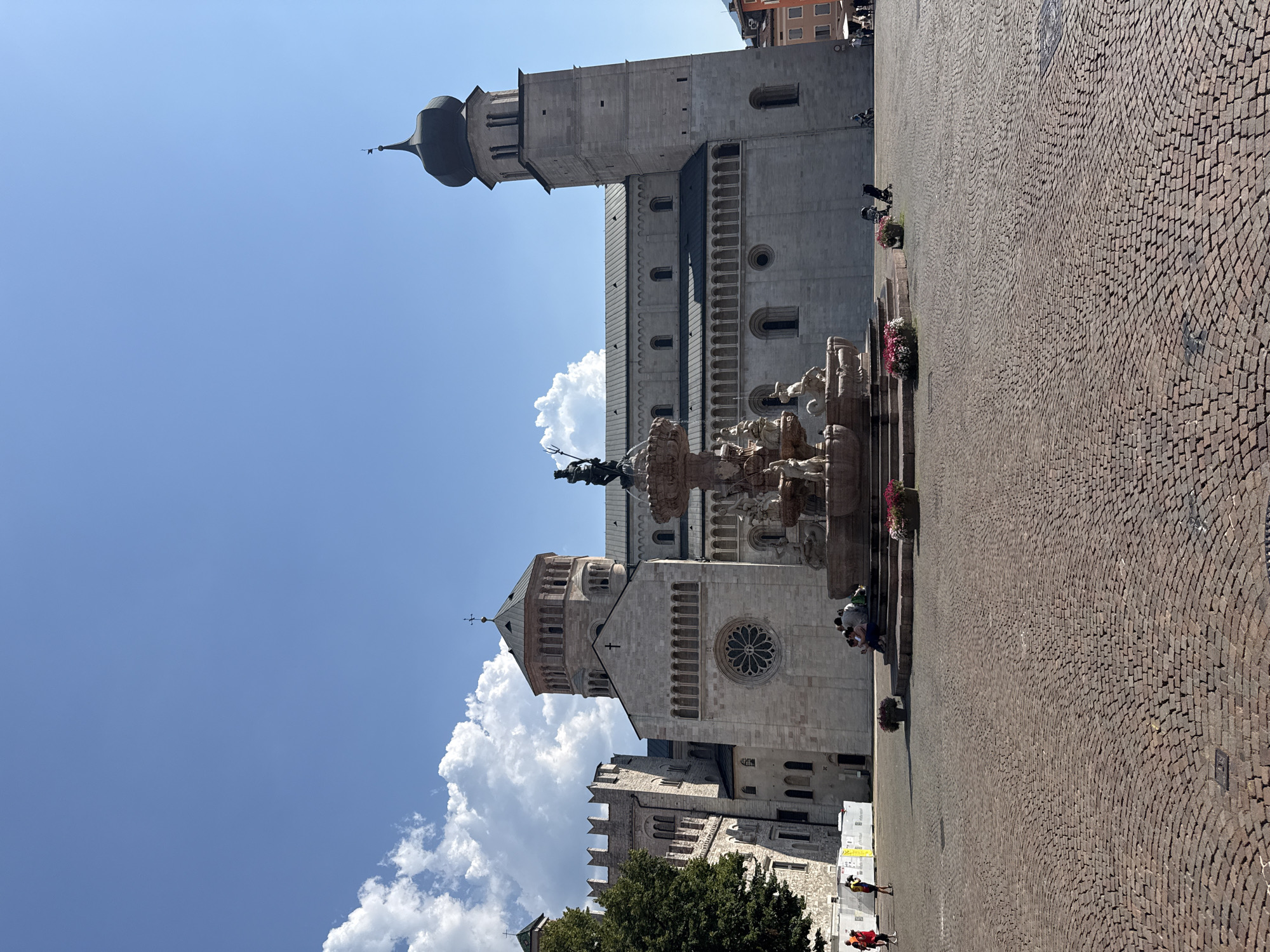 The image shows the Piazza Duomo in Trento, Italy. In the foreground, a cobblestone plaza is visible with several people sitting on the steps of a fountain. The fountain itself is ornate with sculptures and water features. Behind the fountain, the Duomo of Trento, a Romanesque cathedral, stands prominently. The cathedral features two towers, one with a bulbous dome, and a large rose window on its facade. The sky above is clear blue with some fluffy white clouds. The scene is bathed in sunlight, indicating a bright, daytime setting.