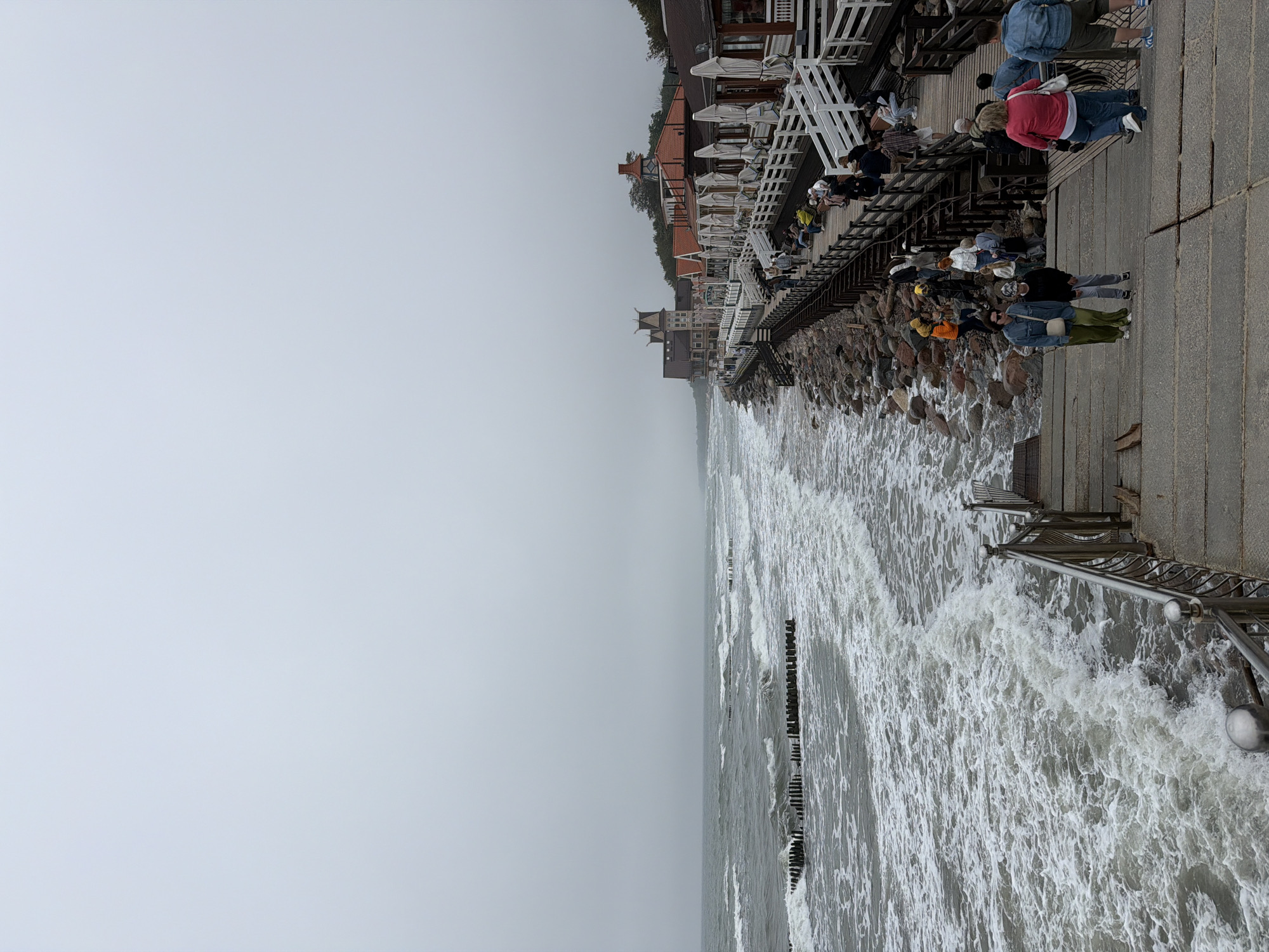 The image shows a view of the Baltic Sea coastline in Zelenogradsk, Russia, under an overcast sky. A wooden boardwalk runs parallel to the shore, lined with buildings that appear to be restaurants or cafes, some with white awnings extended. A stone embankment separates the boardwalk from the sea. Wooden breakwaters are visible in the water. Numerous people are walking on the boardwalk and seated on benches. The foreground features a concrete stairway leading down from the boardwalk. The sea is rough with noticeable waves breaking.