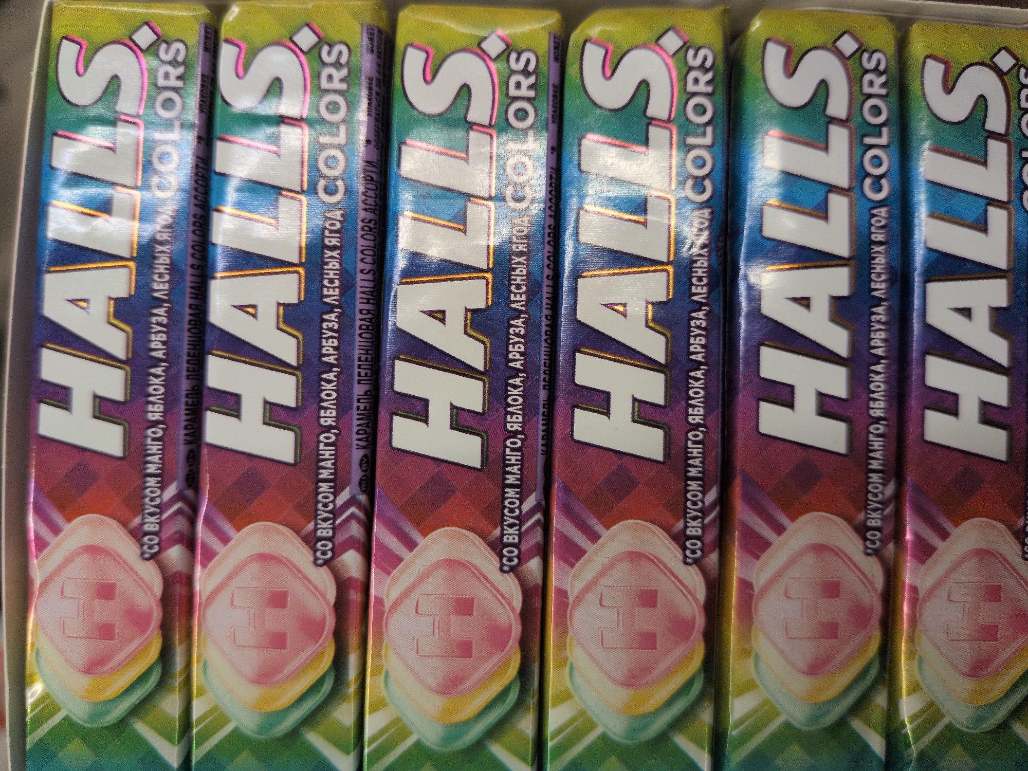Six identical packs of "Halls Colors" lozenges are arranged vertically and closely packed together, likely within a retail display box or tray. The image is a close-up, top-down shot, showing the colorful packaging design prominently. Each pack features the "HALLS COLORS" logo in large, bold letters with a shimmering, iridescent effect. The overall color scheme of the packaging is vibrant, transitioning from yellow and green at the top to blue and purple towards the bottom, with a pattern of diagonal stripes.
The text on the packaging is in Russian, indicating a product sold in a Russian-speaking market, consistent with the location context of Sochi, Russia. Key visible text includes: "СО ВКУСОМ МАНГО, ЯБЛОКА, АРБУЗА, ЛЕСНЫХ ЯГОД" which translates to "WITH THE TASTE OF MANGO, APPLE, WATERMELON, FOREST BERRIES." Below this, it states: "КАРАМЕЛЬ ЛЕДЕНЦОВАЯ HALLS COLORS АССОРТИ," translating to "HALLS COLORS ASSORTED LOZENGES CARAMEL."
Towards the bottom of each pack, an illustration depicts three stacked, translucent lozenges in pink, yellow, and green, each with a stylized "H" imprinted on its surface. The lighting is even, highlighting the glossy finish of the packaging. No people or activities are visible; the focus is solely on the product display.