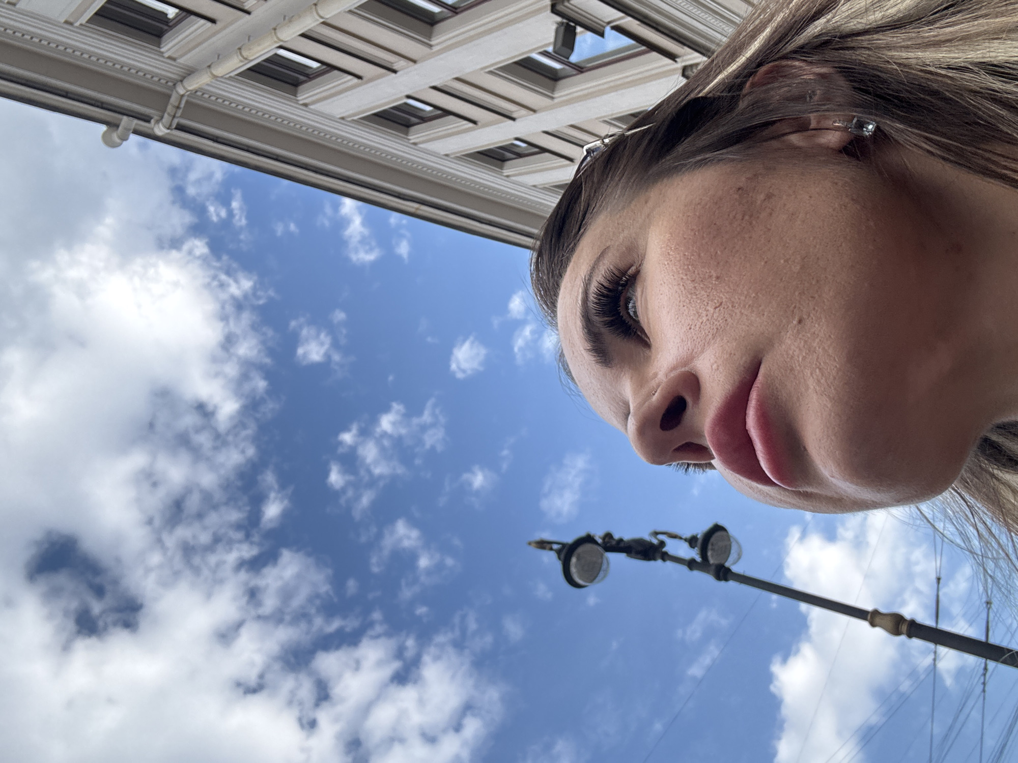 An extreme low-angle perspective shot looks up towards a clear blue sky dotted with white clouds, a classical building facade, and a street lamp, with a person's face occupying the right side of the frame.
The primary subject is the right side of a person's face, seen from below and to the side. Visible features include the nose, full lips (closed), long dark eyelashes, a blue eye, and part of the forehead and cheek, which shows natural skin texture. A small, square-shaped stud earring is visible in the earlobe, and strands of light brown hair frame the face. The person appears to be looking slightly upward and away, towards the upper left of the image, with a neutral expression.
In the upper left background, an ornate building facade with classical architectural elements is visible. It features light-colored stone or stucco, intricate moldings, and what appear to be window recesses or decorative panels on its underside. A white pipe or downspout runs along the building's edge. Below the building and to the left of the person's face, a segment of a street light with two luminaires is visible, set on a dark, possibly cast-iron, pole typical of an older urban setting. Thin wires are faintly visible in the distance.
The sky is bright blue with scattered white cumulus clouds, indicating a clear and sunny day, likely midday or early afternoon. The architecture and street furniture align with the characteristics of a historic European city like Saint Petersburg, Russia. There is no visible text in the image.