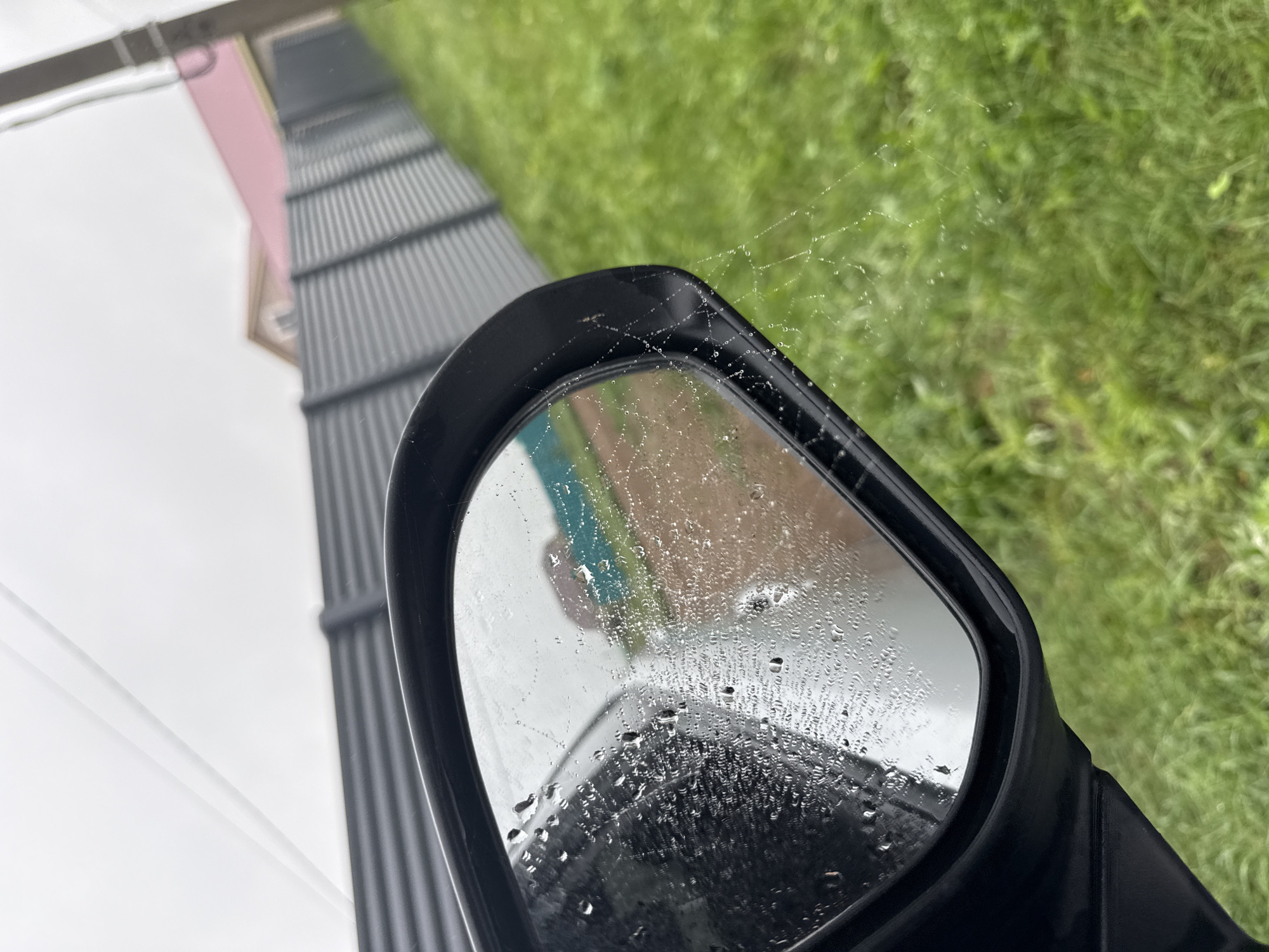 The image shows a car's side mirror reflecting a cloudy sky, a corrugated metal fence, green grass, and part of a house with a pink roof. The mirror is covered in raindrops and a delicate spiderweb with dew drops. The setting appears to be outdoors, likely in a rural or suburban area of Nizhneyarkeyevo, Russia, given the presence of a fence and a house. There are no people visible in the image. The overall mood is somber, possibly due to the overcast weather.