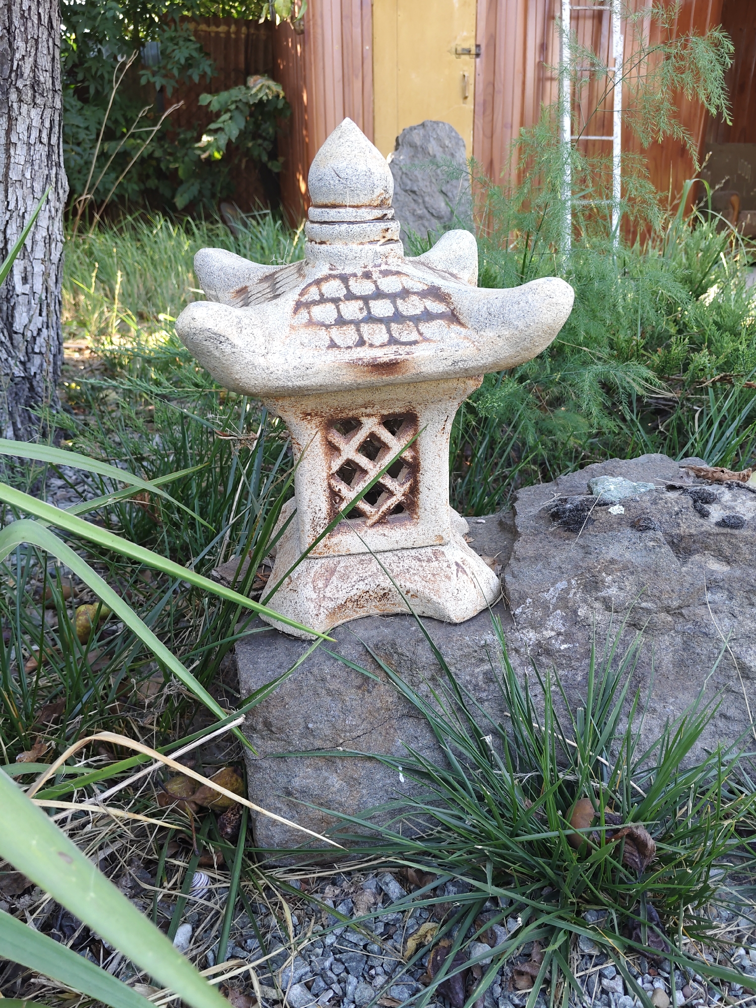 A stone lantern, a typical element of Japanese gardens, is placed on a rock in a garden. The lantern is made of light-colored stone and has a pagoda-like shape with decorative carvings and openings. It is surrounded by green grass, dry leaves, and gravel. In the background, there is a tree trunk, a wooden fence, and some green foliage. The scene is outdoors, likely in a backyard or a park. There are no people or animals visible in the image. The location is not specified as Rostov-on-Don, Russia.