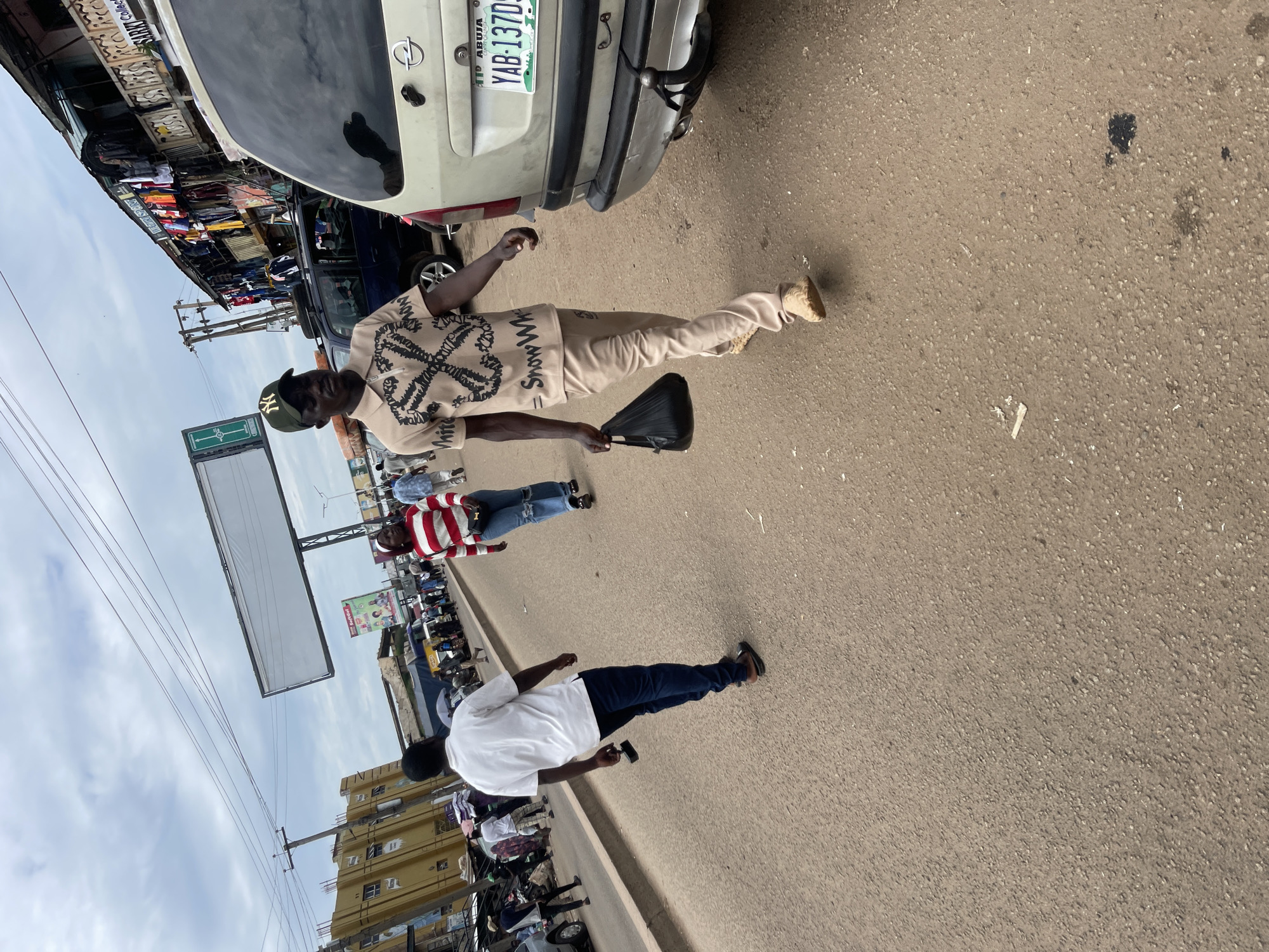The image captures a street scene in Jos, Nigeria, possibly near a market or busy commercial area, characterized by a cloudy sky and a wide asphalt road. In the foreground, a man in a light-colored polo shirt and matching pants, wearing a green baseball cap, walks towards the right, carrying a black plastic bag in his left hand. Behind him and to his left, another man in a white t-shirt and dark pants is also walking away from the camera. Further in the background, several other individuals are seen walking along the road and near the roadside stalls. The roadside is lined with shops displaying various items, including clothing, and a billboard stands prominently above the stalls. A beige Opel car is parked on the right side of the road, its license plate reading "ABUJA YAB-137DS". The overall scene depicts a typical day in a bustling urban environment in Jos.