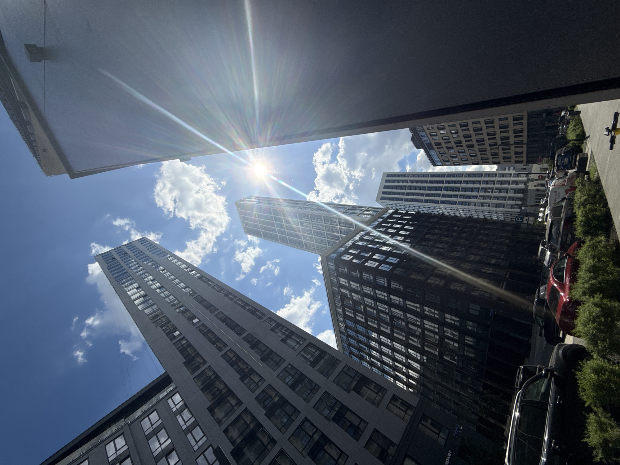 The image presents a low-angle view looking upwards at modern high-rise buildings under a bright, sunny sky with scattered white clouds in Moscow, Russia. The sun is a prominent feature, positioned centrally in the upper sky, emitting strong lens flares and sunbeams.
The scene is dominated by several contemporary, tall buildings characterized by dark grey and black facades and numerous rectangular windows. The perspective suggests the photo was taken from below, possibly from a street level or a lower floor balcony, as a dark, flat overhead structure frames the top of the image and a similar dark structure appears at the bottom.
To the right, a street or parking area is visible, with several cars parked alongside a patch of green bushes. A red car and a black SUV are among those discernible. There is no explicit human activity occurring in the shot.
The clear sky and bright sun indicate it is likely midday or early afternoon. Visible text on a sign on one of the buildings, below the red car, includes the Cyrillic word "маркет" (market) and possibly "Парад" (Parade), confirming the Russian location. The overall impression is one of modern urban architecture on a vibrant, clear day.