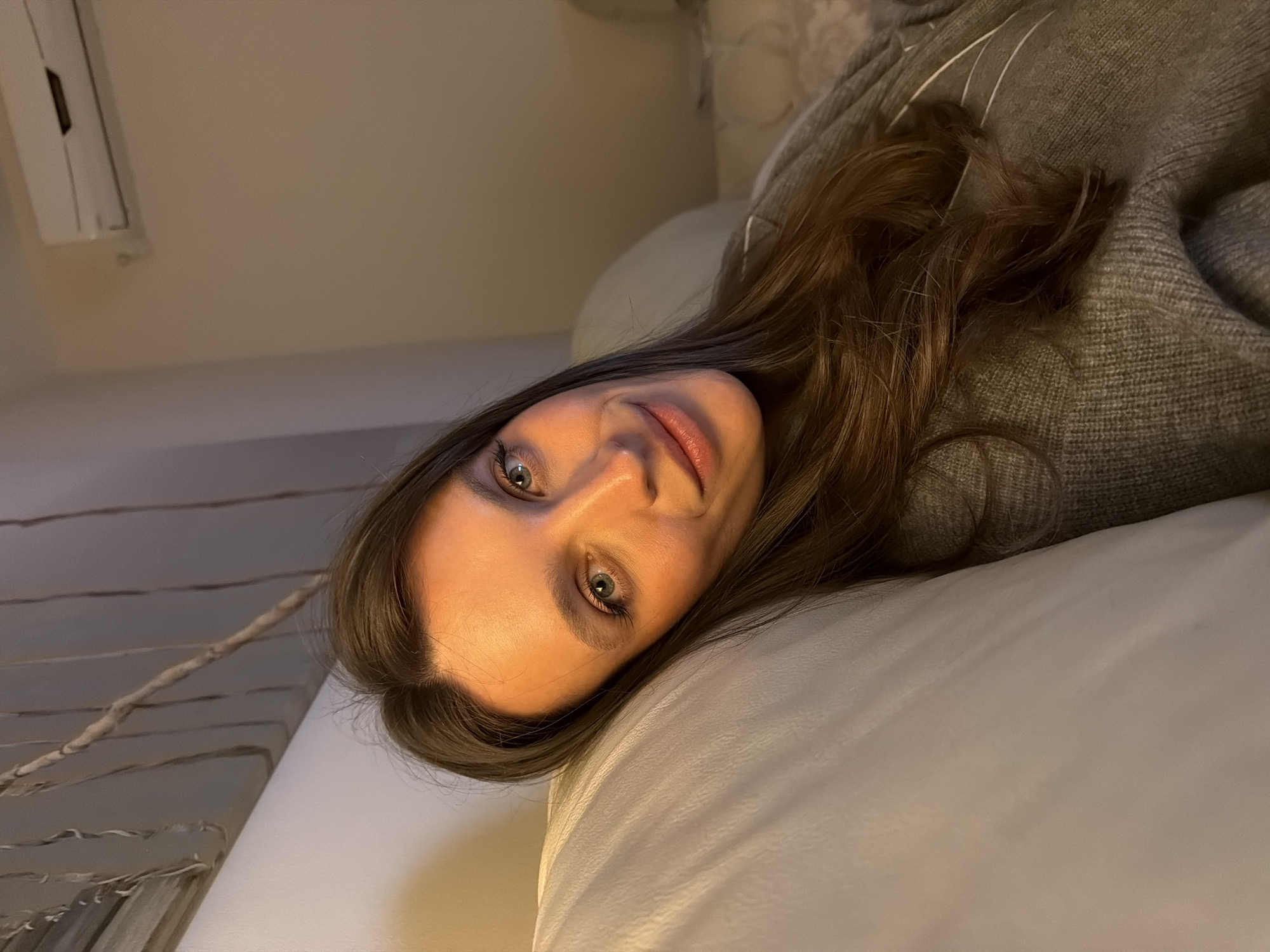 A woman with long brown hair and light eyes is lying on a white surface, possibly a bed or couch. She is wearing a gray sweater. The environment appears to be indoors, likely a residence. The photo doesn't offer sufficient information to locate her precisely in Budva, Montenegro, or to ascertain any specific actions or interactions.