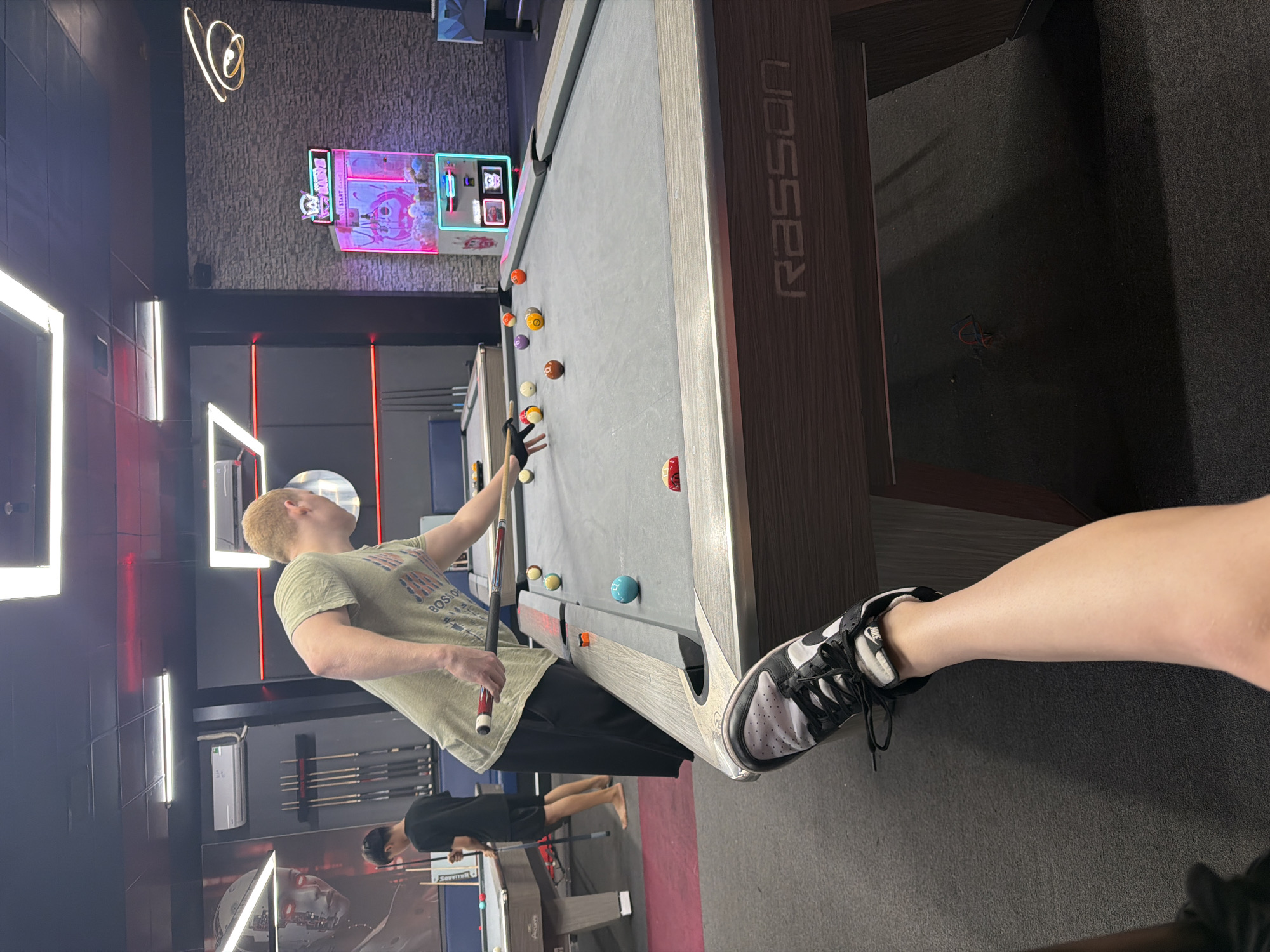 The image depicts an indoor pool hall. In the foreground, a person's leg and foot wearing a black and white Nike sneaker are visible, suggesting the photographer is seated or standing close to a pool table. The main action focuses on another individual, a young man with light-colored hair, wearing a light green t-shirt and dark pants, who is actively playing pool. He is positioned behind a pool table, holding a pool cue and aiming to strike a ball. Several pool balls, including a turquoise one, are scattered across the grey felt surface of the table. In the background, another person, a young male in a black t-shirt and shorts, stands near a pool table, holding a cue. The environment includes overhead lighting fixtures, walls adorned with what appears to be artwork or decorative elements, and a "claw" arcade machine. The overall scene is that of a recreational establishment. The provided location "Bac Nha Trang Ward, Vietnam" is noted, though there are no specific visual cues in the image that directly confirm this geographical location.