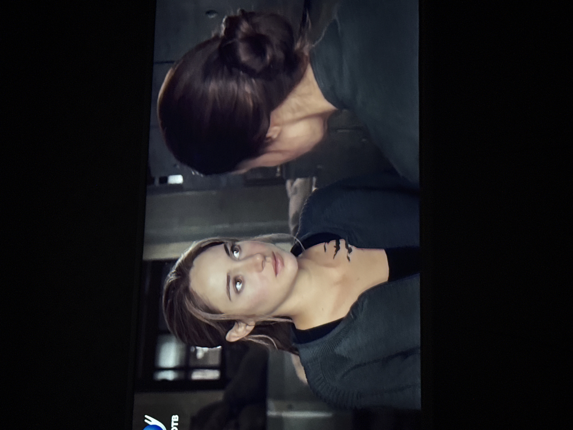 Two women are present in the scene. One woman lies on her back with a tattoo on her neck. The other woman, with her hair in a bun, is leaning over her. The environment is not clearly discernible, but it seems to be an interior space. It is not possible to determine any location including the city of Mytishchi, Russia, from this image.