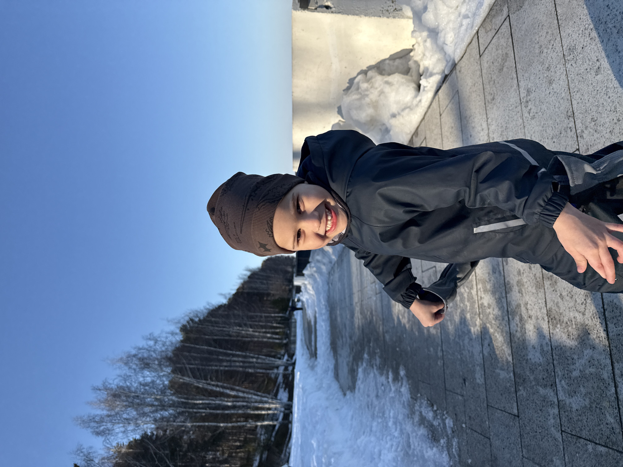 A young child wearing a brown beanie and a dark jacket is standing on a paved walkway in Chaikovsky, Russia. To the left, bare trees climb a hill under a clear blue sky. Patches of snow line the walkway and are visible at the edge of the concrete wall in the background. The child is smiling and appears to be outdoors on a sunny day.