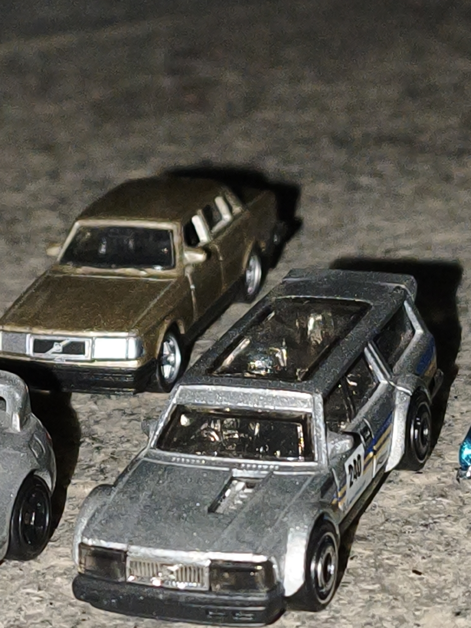 On a dark, textured surface, likely concrete, are several toy cars. A metallic grey Volvo 240 race car with the number 230 prominently displayed is in the foreground, featuring a modified hood and roll cage visible through the open roof. Behind and to the left of it is a gold-colored Volvo 240 sedan. Partially visible on the left edge of the frame is another grey toy car, and a blue toy car is glimpsed on the right. The setting appears to be outdoors, likely at night, given the lighting and background.