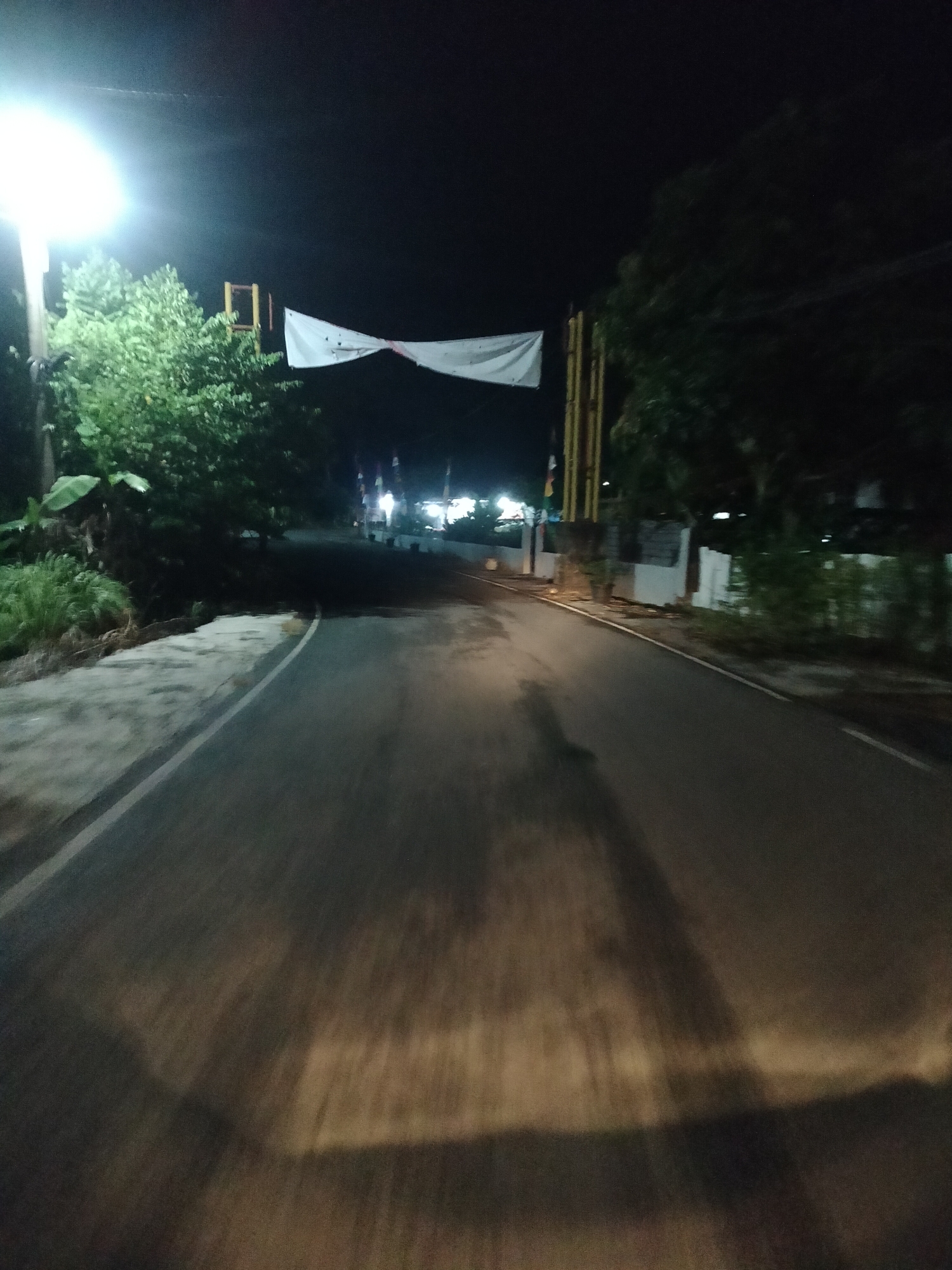 This is a nighttime, first-person perspective photograph of a road in Kumbangindah, Indonesia. The road is paved and appears to be a two-lane street. On the left side of the road, there is a dirt shoulder with grass and some banana trees. Further back, lush green trees line the area. A bright street light illuminates the scene from the left. A white banner with an unknown design is stretched across the road in the distance, supported by yellow poles. On the right side of the road, there are some buildings with white walls and green foliage in front of them. The road surface is wet and reflects the lights. There are visible shadows on the road, suggesting movement. The overall atmosphere is dark and quiet.