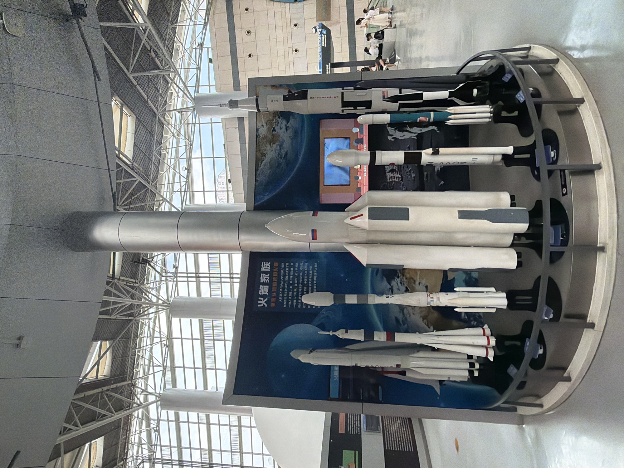 The image depicts an indoor exhibition showcasing a collection of model rockets. The models are arranged in a circular display, with a large, central pillar dominating the foreground. A curved, dark railing encloses the models. Behind the rockets is a large display board with Chinese text and an image of Earth from space. To the right of the display, several people, including adults and children, are observing the exhibits. The background reveals the architecture of a modern building, with a high ceiling supported by metal trusses and large windows or skylights. The flooring is a light-colored, smooth surface. Based on the context of the exhibition and the commonality of such displays in scientific museums, this scene is likely located in a science museum within 江北城街道, China.