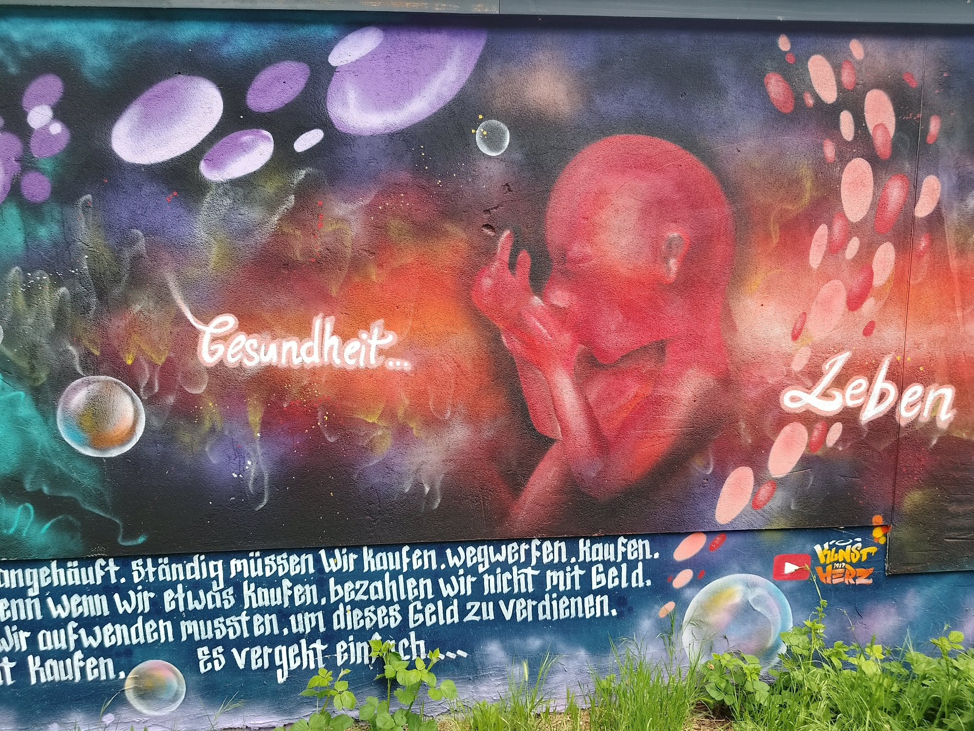 A mural painted on a rough, uneven concrete wall depicts a fetus-like figure rendered in various shades of red and crimson. The background consists of blurred, nebulous shapes in shades of purple, orange, and black, interspersed with floating bubbles. The words "Gesundheit..." and "Leben" are written in white script on either side of the fetus. Below the mural, a section of the wall is painted in a darker color, with white text that reads, "angehäuft. Ständig müssen Wir kaufen.wegwerfen.Kaufen. enn wenn wir etwas Kaufen, bezahlen wir nicht mit Geld. Wir aufwenden mussten, um dieses Geld zu verdienen. it Kaufen. Es vergeht ein ch..." To the right, there is a small logo or tag with the text "KUNST MIT HERZ." Grass and small green plants grow at the base of the wall, partially obscuring the lower section.