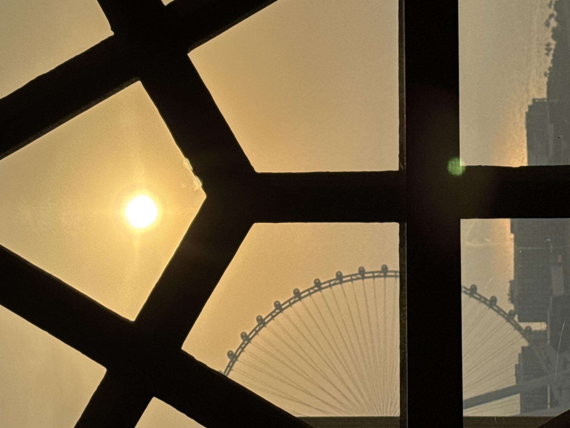 The image captures a view through a window with a dark, geometric frame. Through the frame, a bright sun is visible in the hazy sky, casting a warm, orange glow. To the left of the sun, a large Ferris wheel is partially visible, appearing as a silhouette against the bright sky. The spokes of the Ferris wheel radiate outwards. Below, in the distance, a cityscape with buildings and palm trees is faintly discernible along the waterfront. The scene suggests an urban environment during either sunrise or sunset, common in Dubai, United Arab Emirates. The presence of the Ferris wheel strongly suggests a location near Ain Dubai or a similar attraction in the city.