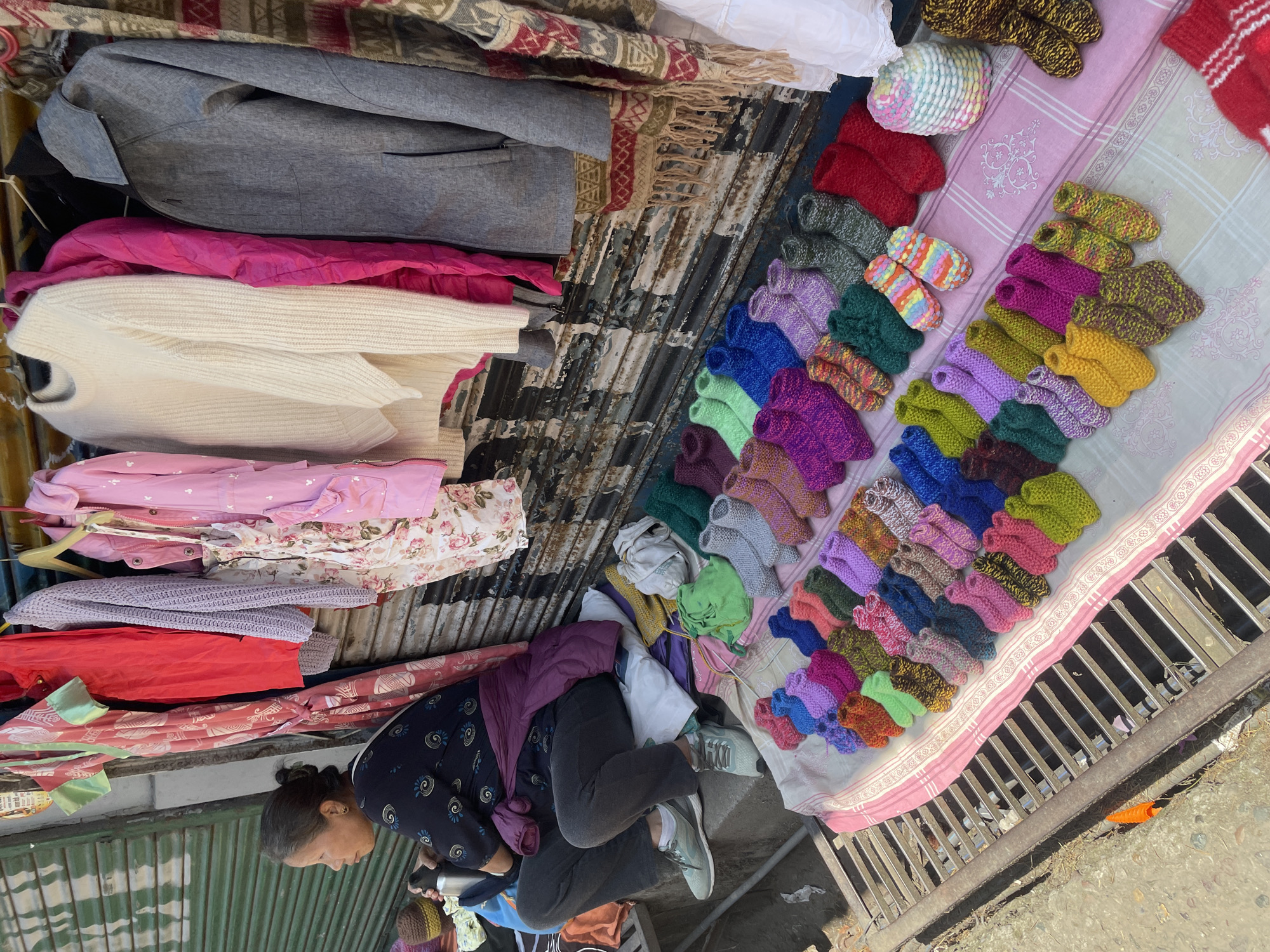 A vendor sits beside clothing on display at a roadside stall in Dharamshala, India. The woman is kneeling wearing a long-sleeved patterned top, a purple shawl, grey trousers, and light-colored sneakers. She is looking down with her hands near a thermos. To her left is a stack of folded clothes in varying colors. Above the clothes is corrugated metal, and above that, more displayed clothing, including a grey blazer and a patterned scarf. To her right, on a pink striped cloth, is a display of hand-knit socks in many colors. The stall is situated on a metal grated structure.
