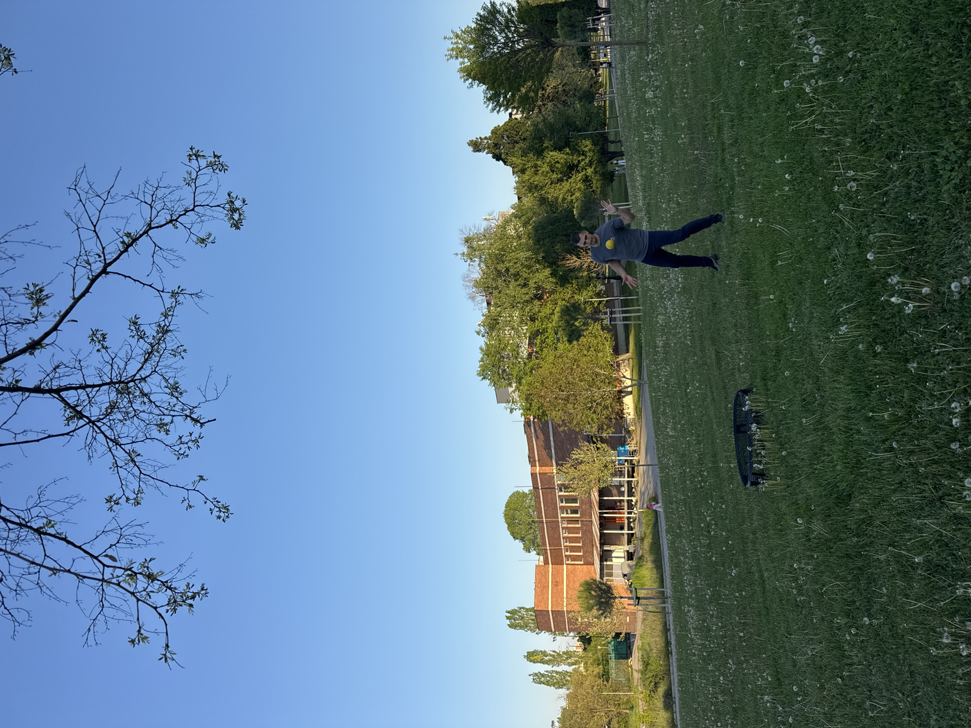 A man is jumping in a grassy park area. The park is located near a brick building. The sky is clear and blue. A bare tree branch is visible in the upper left corner. Sofia, Bulgaria is not identifiable as the location from the available details.
