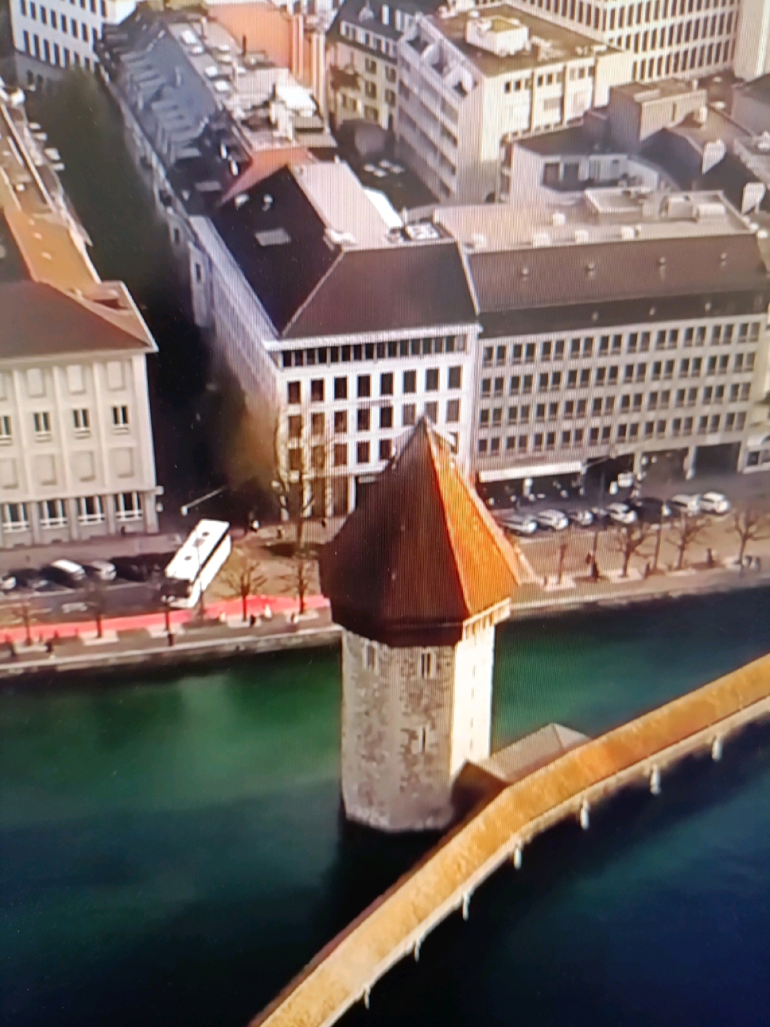 An aerial, high-angle, medium shot captures a segment of a city on a sunny day. In the foreground, a body of green water is visible, with a light brown, arched bridge extending across it from the bottom right corner towards the center. Partially submerged in the water and adjacent to the bridge is a hexagonal, stone tower with a reddish-brown, conical roof. Behind the tower, on the far bank of the water, is a street lined with trees and vehicles, including a white bus. Beyond the street, several multi-story buildings with light-colored facades and dark roofs are clustered together, forming a cityscape. The architecture suggests a European style. The image does not depict anything that would suggest the location is Netanya, Israel.