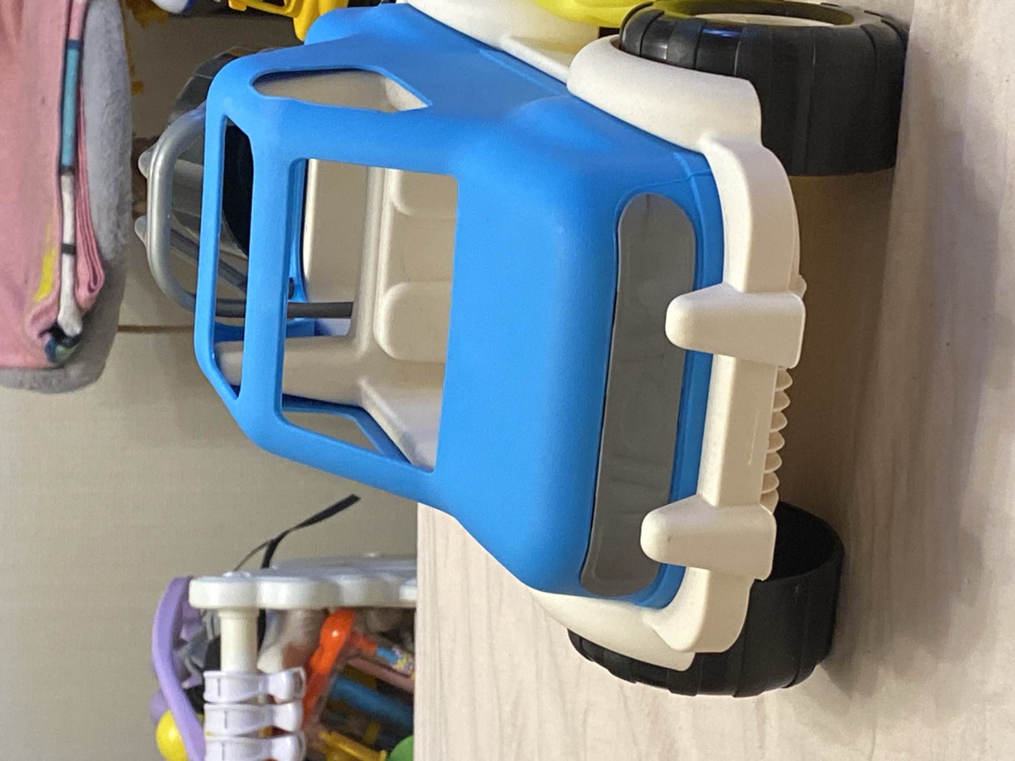 The image shows a close-up, eye-level view of a blue and white toy truck. The truck is the primary subject, positioned centrally in the frame, with its front facing forward. The toy has black wheels with thick treads. The body of the truck is predominantly blue on the top and hood area, with a white chassis and bumper. A gray rectangular detail is present on the front of the truck, resembling a grille or headlight assembly.
In the background, to the left of the truck, is a blurry white plastic toy structure with several white pegs or clips on it. Further back and to the left, there are hints of colorful toys, including a yellow ball and some orange and blue objects. Above the truck and slightly to the right, a portion of a gray and pink fabric item is visible, possibly a blanket or stuffed toy. The background wall appears to be a neutral color. The truck is resting on a light-colored surface, likely a table or floor, with a soft, textured appearance. The lighting suggests an indoor environment, possibly illuminated by overhead light. The image is static and does not depict any action or people.