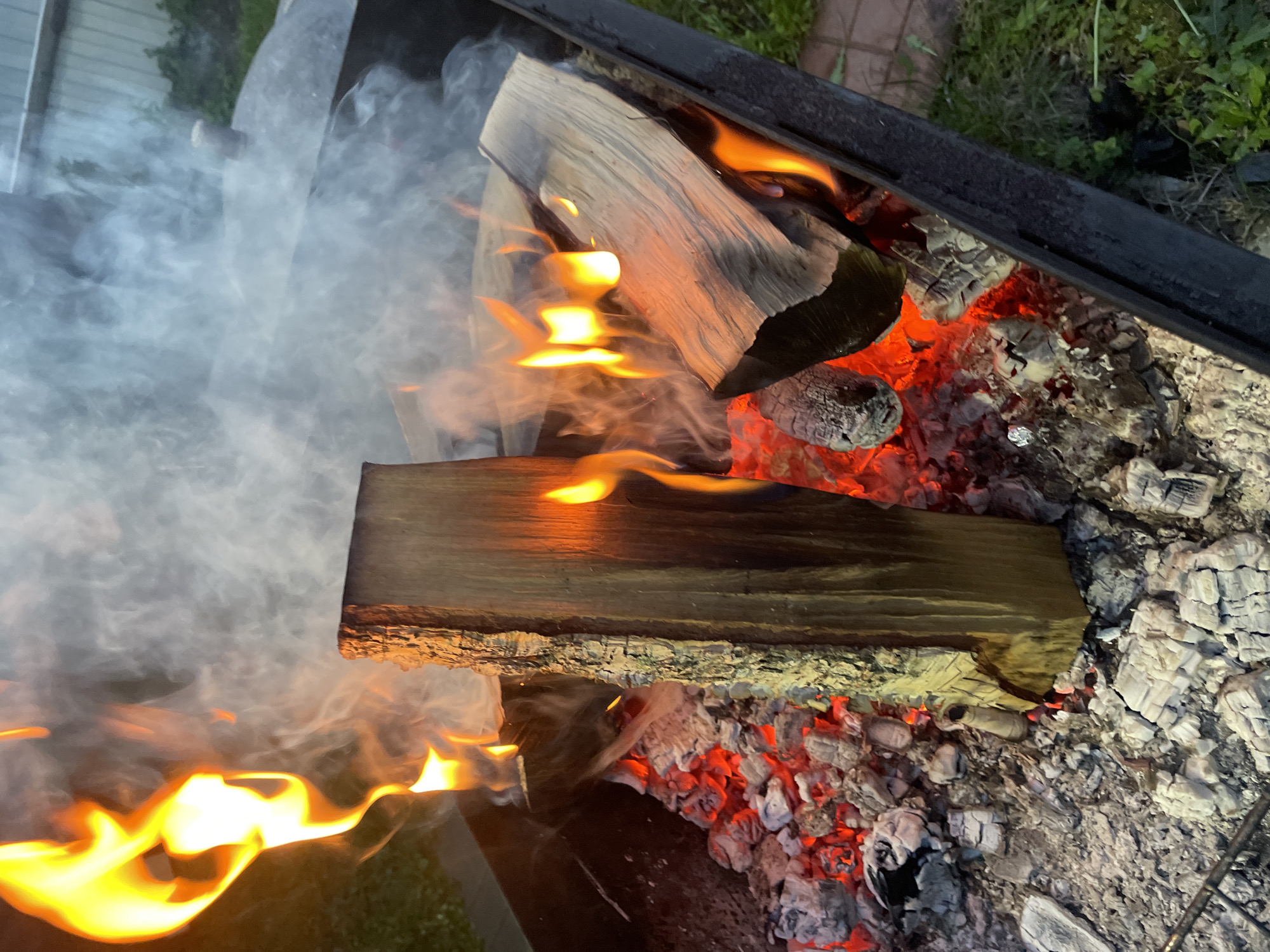 The image shows a fire in a black metal fire pit or grill. Two large pieces of wood are burning with flames and smoke. Hot coals and ash are visible beneath and around the wood. The scene appears to be outdoors. Some vegetation is visible in the background. There is no indication of the location being Bibikha, Russia, or any specific location. No people are visible.