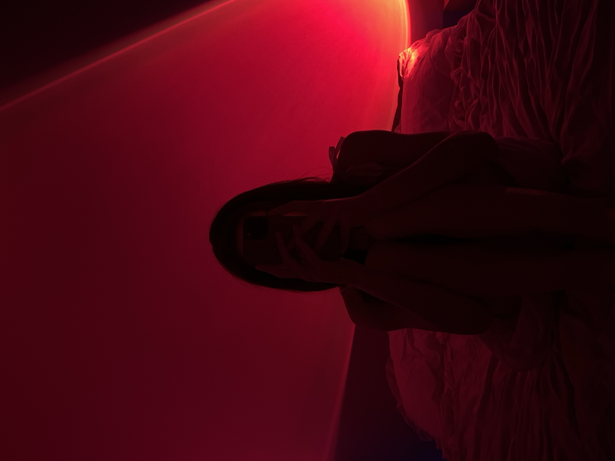 A person is sitting on a bed covered with crumpled white sheets. They are holding a dark-colored smartphone in front of their face, obscuring it from view. The person's body is partially silhouetted against a red background, which is illuminated by a light source casting a gradient effect. The environment appears to be a bedroom, with a pillow visible behind the person. The image was taken in Ternopil, Ukraine.