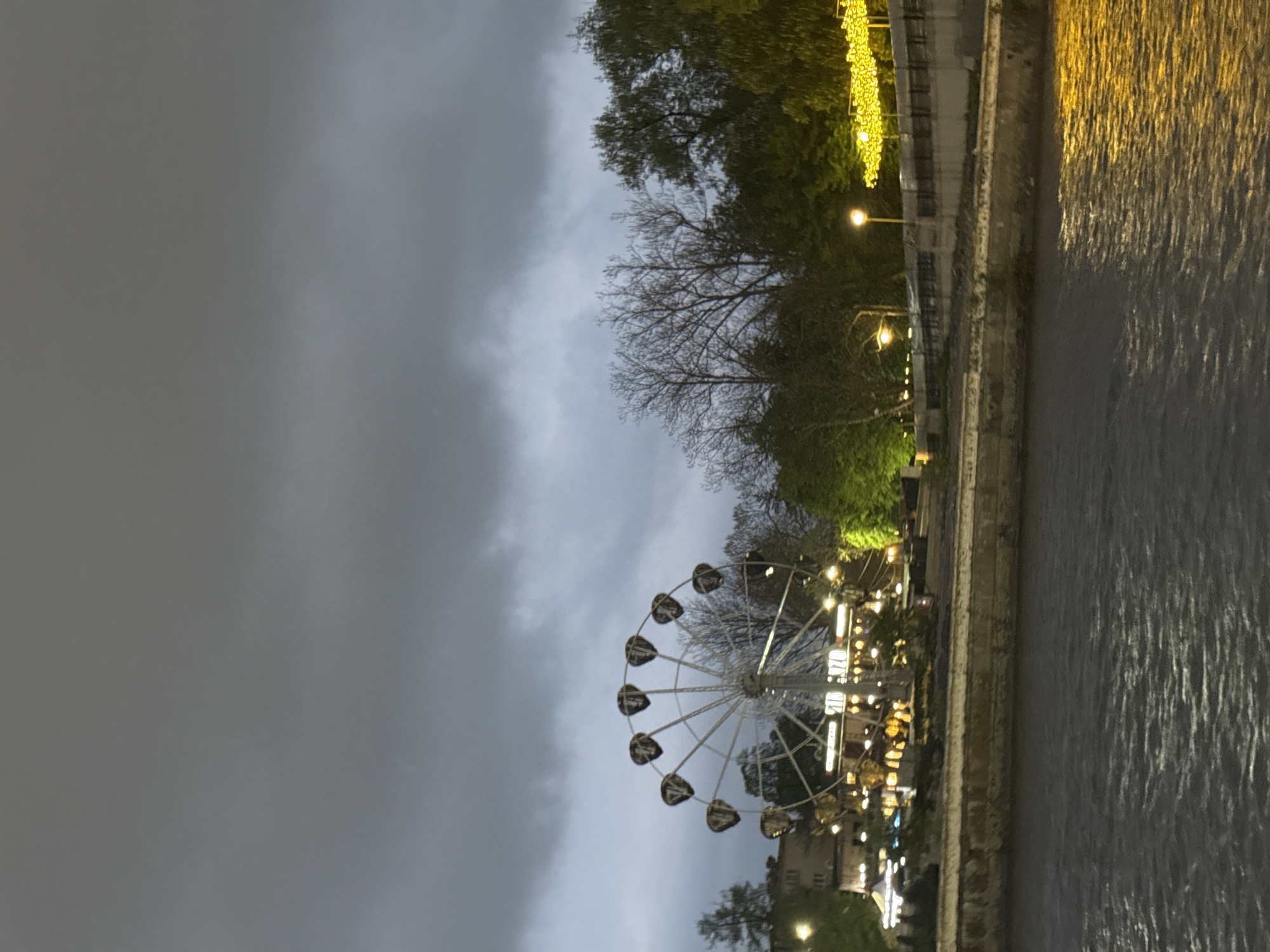 The image depicts a Ferris wheel against a cloudy sky, likely at dusk or night. The wheel is illuminated, and its carriages appear dark. Alongside the Ferris wheel, there is a river or body of water, with light reflecting off the surface, suggesting illumination from nearby structures. The opposite bank is lined with trees and buildings. Lights are strung along the edge of the river, and the scene seems to be in an urban environment. Although the city name Komgaron and its location in Russia is mentioned, there's nothing in the image to explicitly confirm this specific location.