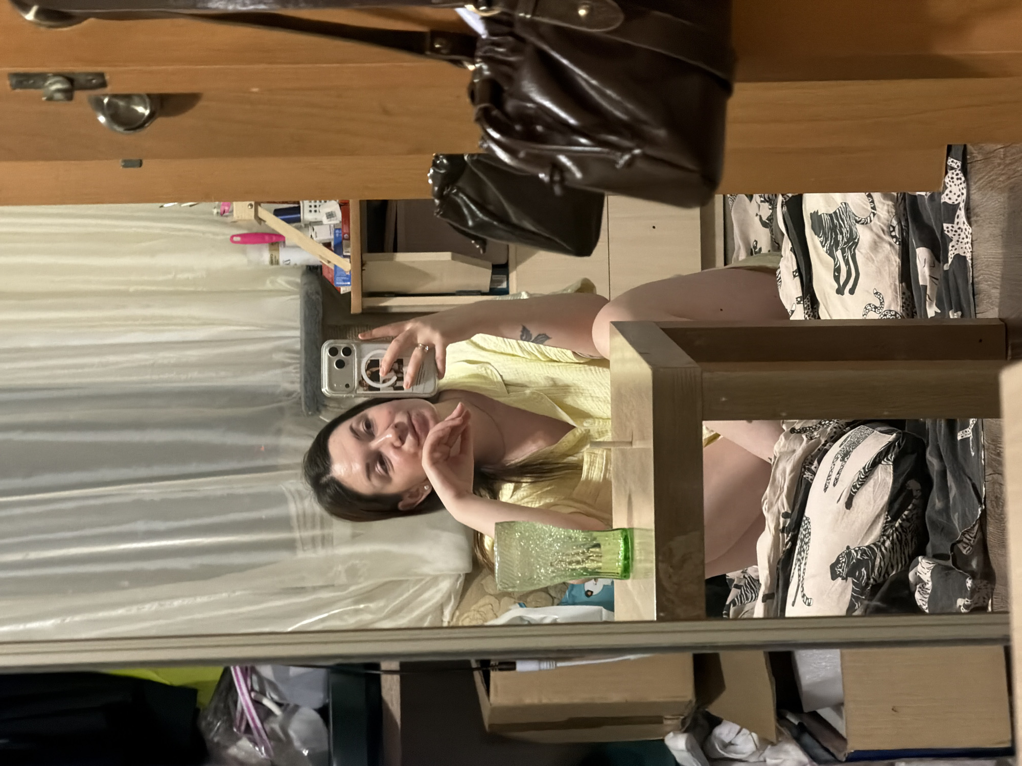 The image shows a woman taking a selfie in a mirror. The mirror is positioned near a wooden dresser with a brown leather purse hanging on it. The woman is wearing a yellow shirt and has dark hair. Her reflection shows that she is holding a smartphone to take the picture. On the shelf behind her, there is a glass and other household items. The shelf also has fabric with animal prints on it. The location or connection to Izhevsk, Russia cannot be determined.