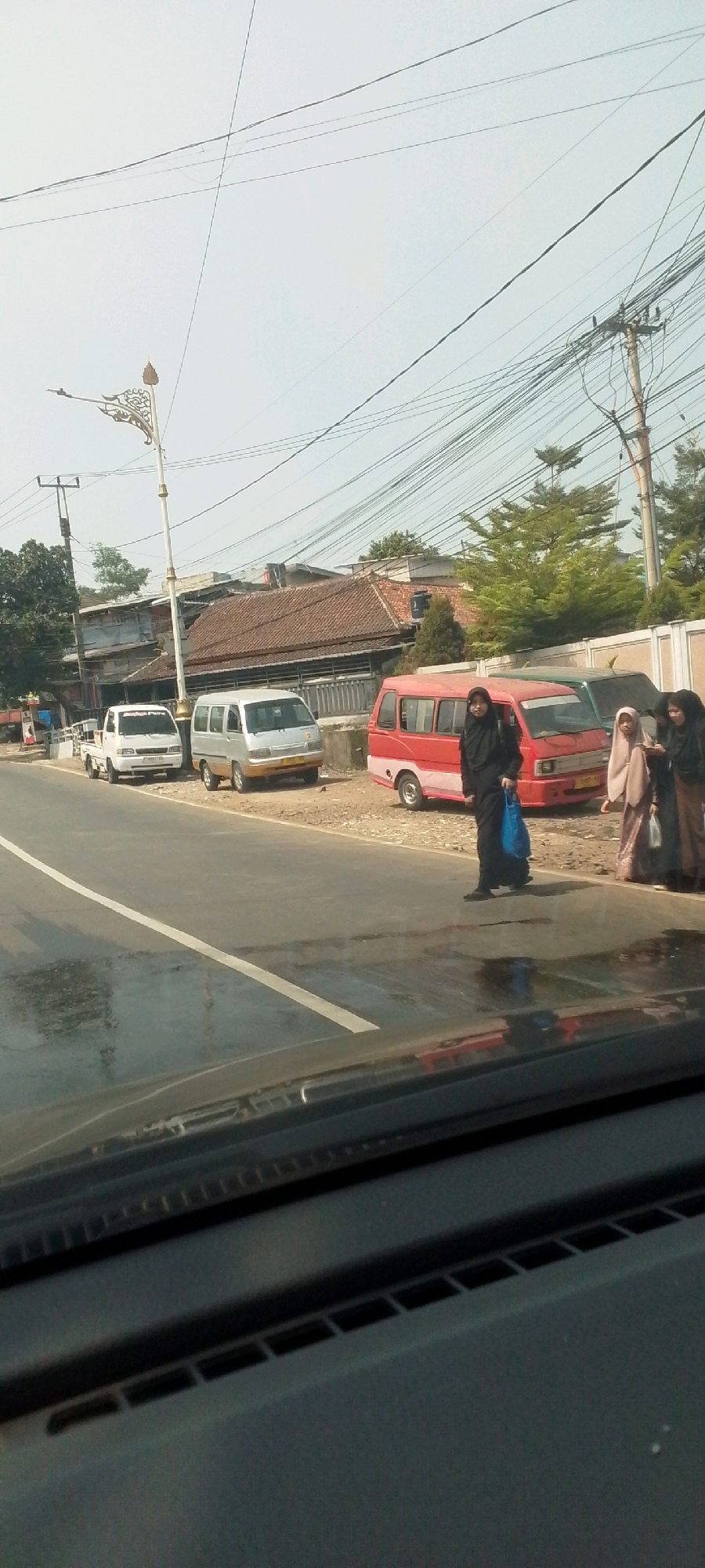 The image depicts a street scene in Cilaku, Indonesia, with a focus on several vehicles and pedestrians. On the right side of the road, a woman wearing a black hijab and a dark abaya is walking across the street, carrying a bright blue bag. To her right, two other women are standing and observing. Behind them, a red and white van is parked. Further down the road, a silver minivan and a white pickup truck are also parked. The road surface appears wet, suggesting recent rain. Overhead, a complex network of power lines crisscrosses the sky, supported by wooden poles. Buildings with terracotta roofs are visible in the background. The scene is captured from the perspective of someone inside a vehicle, with a portion of the car's dashboard visible in the foreground.