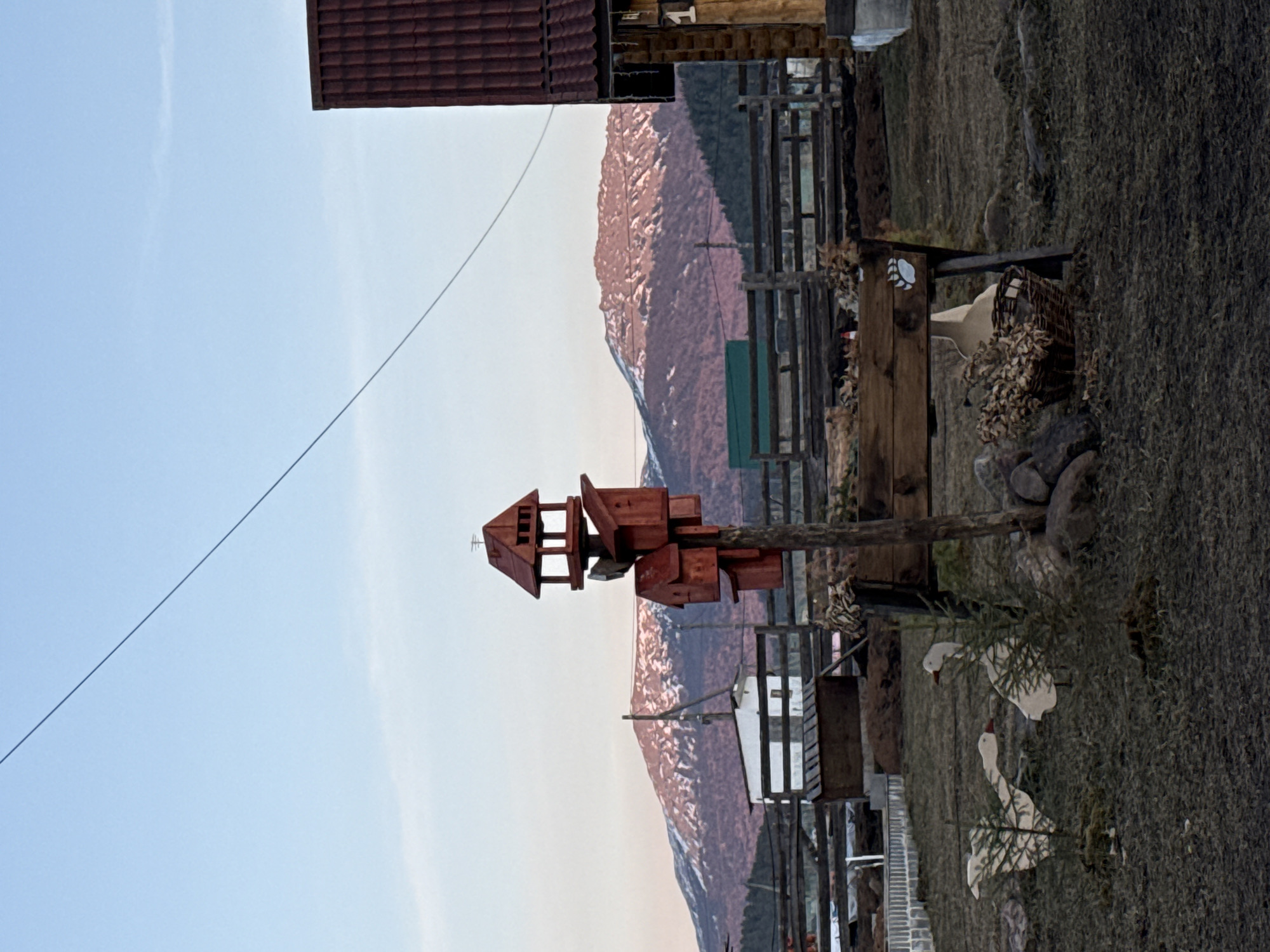 The image captures a scene in Tyulyuk, Russia, featuring a wooden multi-tiered birdhouse prominently placed in the foreground. The birdhouse stands against a backdrop of a mountain range with snow-capped peaks. In the immediate foreground, the ground is covered with grass and scattered with decorative wooden geese. A fence runs parallel to the mountain range, partially obscured by the birdhouse. A wooden structure with a basket and various other objects is situated near the fence. The sky above is a gradient of light blue.