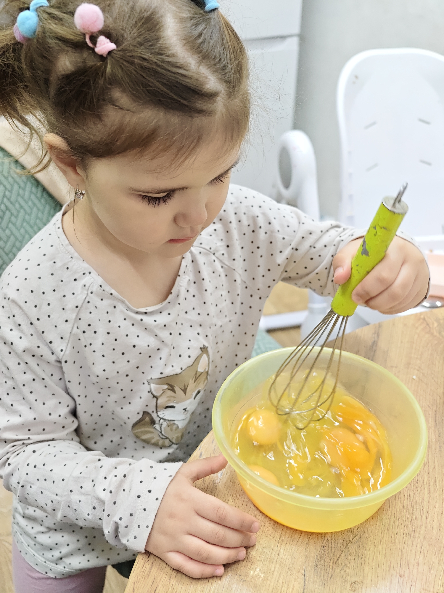 A young girl with brown hair styled in pigtails with colorful hair ties is beating eggs in a yellow bowl with a green whisk. She is wearing a long-sleeved, light-colored shirt with black polka dots and a cartoon fox on it. The bowl is on a wooden table. The background includes a green cushion, a white baby chair, and a white refrigerator. The photo was likely taken in a kitchen in the city of Chaikovsky, Russia.