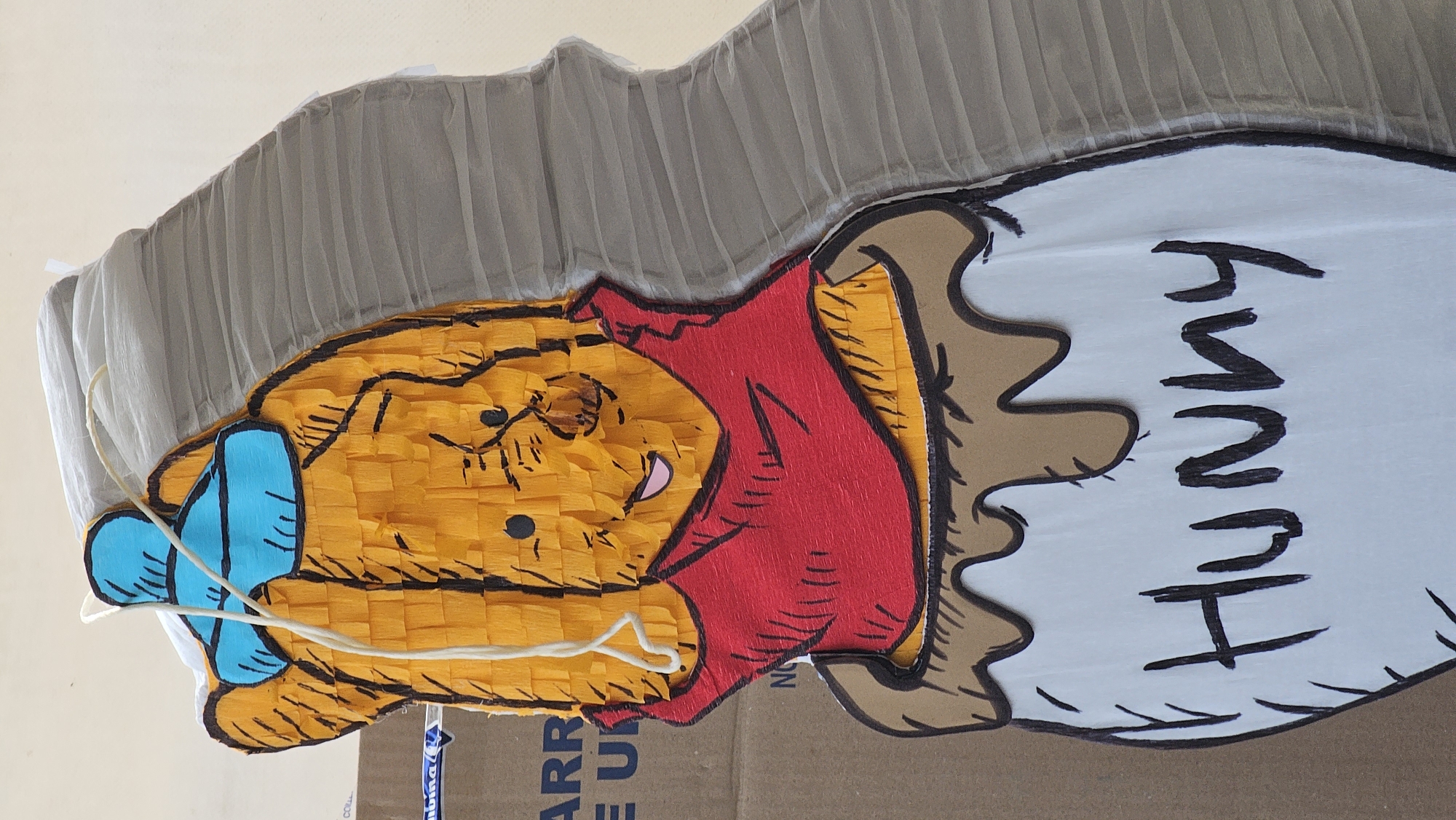 The image shows a piñata depicting Winnie the Pooh holding a jar of "Hunny". The piñata is predominantly orange with red clothing and a blue bow on the hand. The "Hunny" jar is white with brown edging and black lettering. The piñata appears to be made of tissue paper and cardboard. The background is a cardboard box and a white, textured surface, possibly fabric. The location is not discernible from the image, and there's no way to connect the piñata with the city of San Bernardino Tlaxcalancingo, Mexico based on the available information.