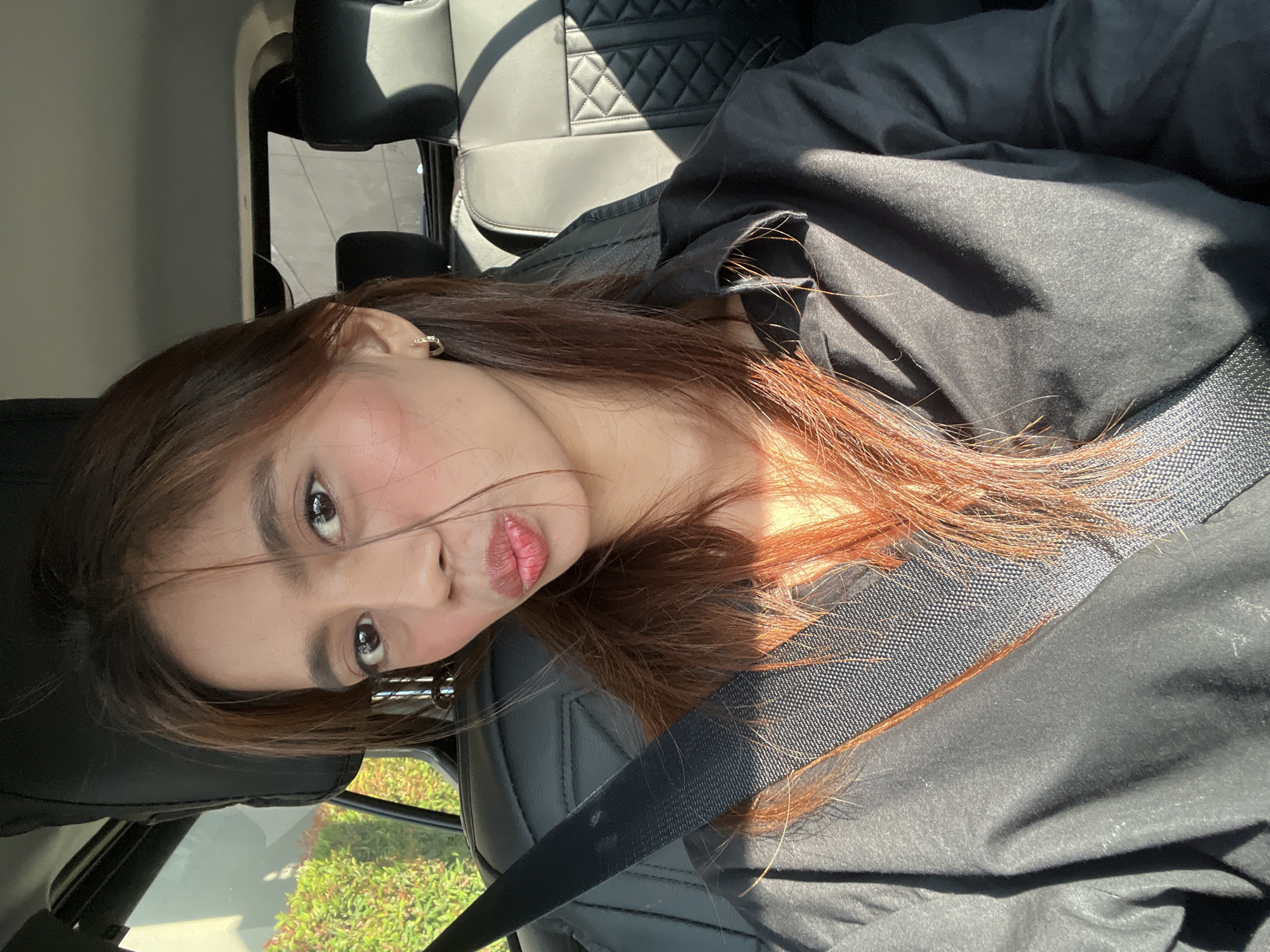 A young woman is depicted in a front-facing photograph, seated in the driver's or front passenger's seat of a vehicle in Davao City, Philippines. She has long, reddish-brown hair with some strands falling across her face. Her dark brown eyes are directed towards the camera, and her lips are pursed in a slight pout, accented with pinkish-red lipstick and visible blush on her cheeks. A small, gold-colored hoop earring is visible on her right ear. She wears a dark, possibly black, collared shirt or jacket, with a black seatbelt securely fastened across her chest from her right shoulder to her left hip.
The car's interior features black leather seats with a diamond-quilted pattern visible on the rear seat upholstery. Bright sunlight enters the vehicle from the right, casting sharp shadows on the woman's clothing and the car's interior, and illuminating sections of her hair with an orange hue. Through the left window, vibrant green foliage and shrubbery are visible, suggesting an outdoor, possibly suburban setting. The right-side window reveals a blurry, light-colored building or structure in the distant background.