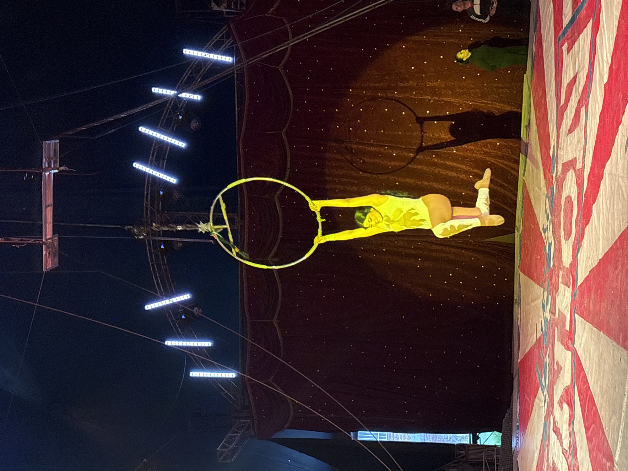 A female acrobat is performing inside a circus tent in Tejalpa, Mexico. She is holding onto an aerial hoop, dressed in a yellow leotard and white boots. The tent interior is visible, including a red and white striped wall and lighting fixtures. There are also dark spots on the circus tent. There are people at the top of the picture, but they are not the main focus of the picture.