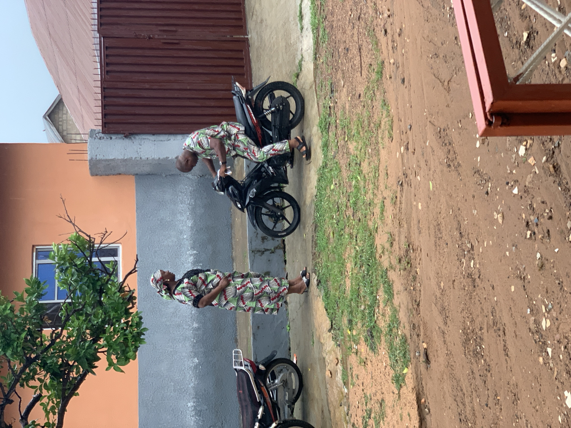 In Kabba, Nigeria, the image captures a daytime scene. A man in patterned clothing is crouched over a black motorcycle, possibly examining or working on it. Nearby, a woman, also dressed in patterned attire, stands on a paved surface. Another motorcycle is parked in the vicinity. The setting includes buildings with a mix of orange and grey walls. The ground transitions from pavement to a dirt and grassy area. The image's right side features a partial view of a brown metal structure. A tree with green foliage is present on the left side of the image.