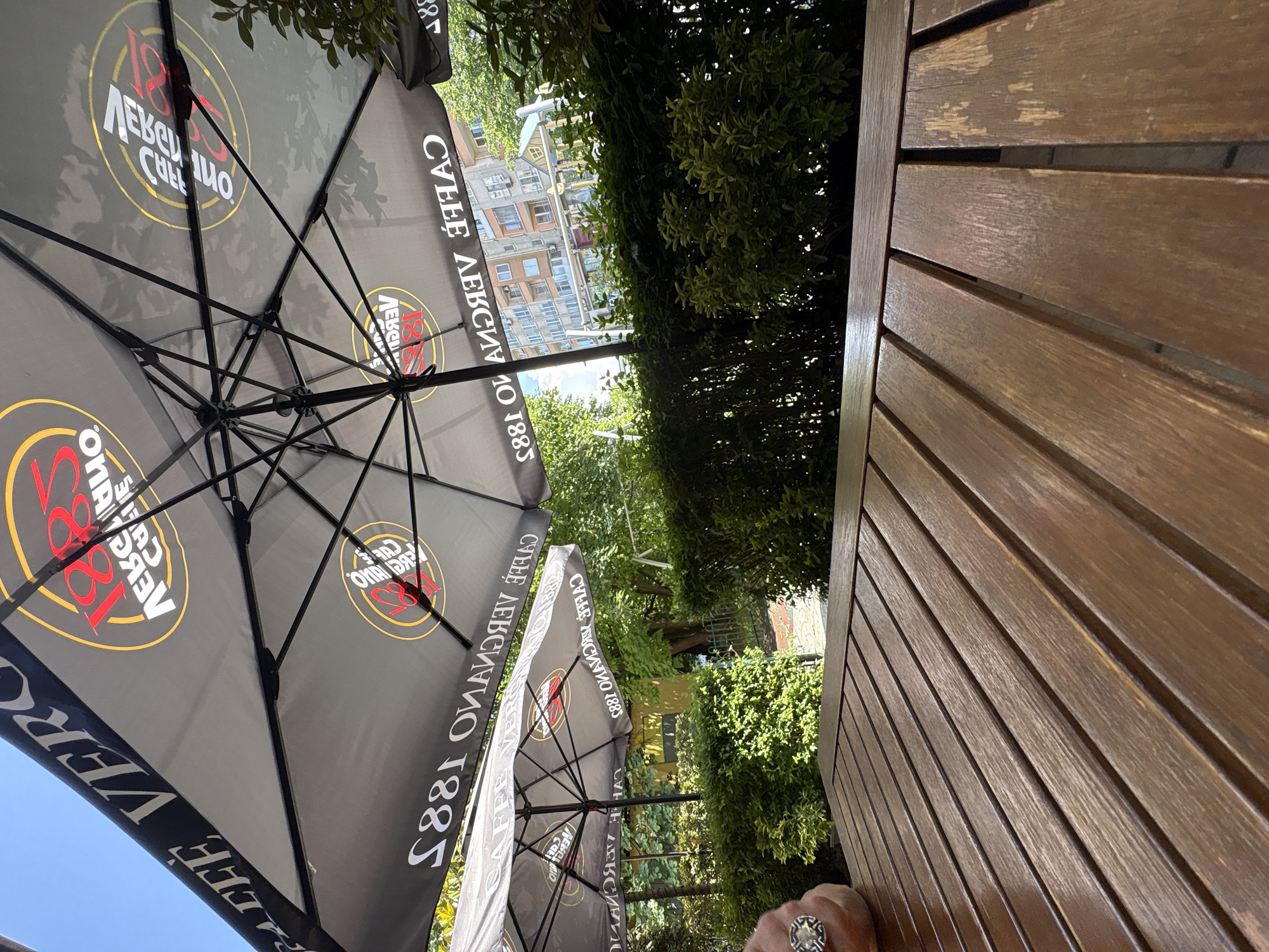 The image, rotated 90 degrees clockwise, captures an outdoor cafe setting on a bright, sunny day, likely in Burgas, Bulgaria. The primary subjects are several large, dark grey patio umbrellas, prominently featuring branding, and a section of a light brown wooden slatted table or chair back in the foreground.
The umbrellas are the dominant feature, displaying text and a logo. The logo appears to be a circle with "1882" in red and what looks like "VERO CAFFE" or "CAFFEE VERGNANO" around it, indicating a coffee brand. The text is visible multiple times on the different umbrella segments.
Behind the umbrellas and green foliage, multi-story residential buildings with balconies are visible under a clear, bright sky. Lush green trees and bushes create a natural screen between the cafe area and the buildings. In the lower right corner, a hand wearing a distinctive ring with a circular, possibly metallic or stone, design is briefly visible, resting on the wooden surface.
The wooden surface, likely part of an outdoor table or chair, shows some wear and tear with areas of lighter wood visible through the finish, suggesting it's well-used outdoor furniture. The scene evokes a relaxed atmosphere, typical of an outdoor cafe or restaurant patio.