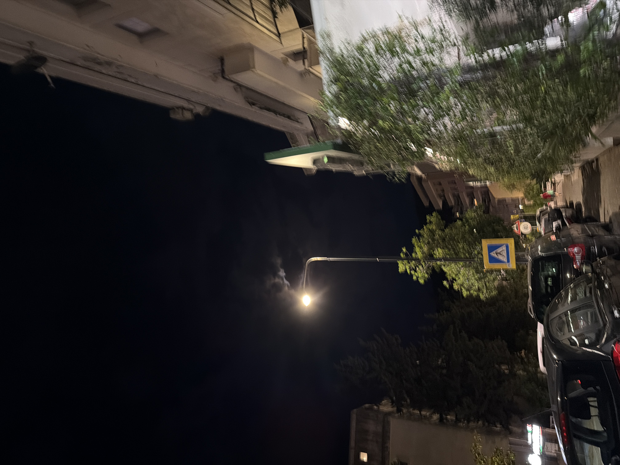 The image depicts a street scene at night in Budva, Montenegro. A street lamp illuminates the street and a pedestrian crossing sign. Several cars are parked along the street, and trees line the sidewalk. In the background, buildings are visible. The sky is dark, with some clouds and a faint light, possibly from the moon. The overall atmosphere is quiet and peaceful.