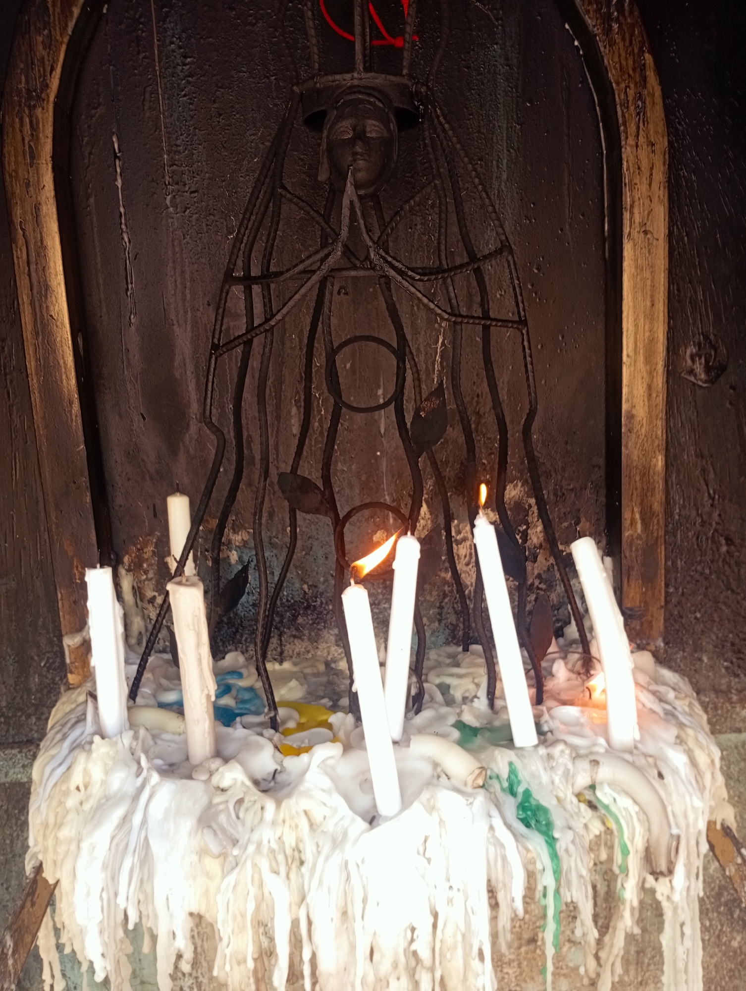 The image depicts a religious shrine or niche, likely located in El Valle del Espíritu Santo, Venezuela, given the context. The shrine features a dark metal sculpture of a praying figure, possibly the Virgin Mary, enclosed within a wooden frame. The base of the shrine is covered in a thick accumulation of melted candle wax, with several lit candles interspersed among the solidified wax. The melted wax shows streaks of white, yellow, blue, and green colors. The background appears to be a dark, possibly stone or concrete wall. The scene conveys a sense of devotion and tradition, with the candles representing offerings or prayers.