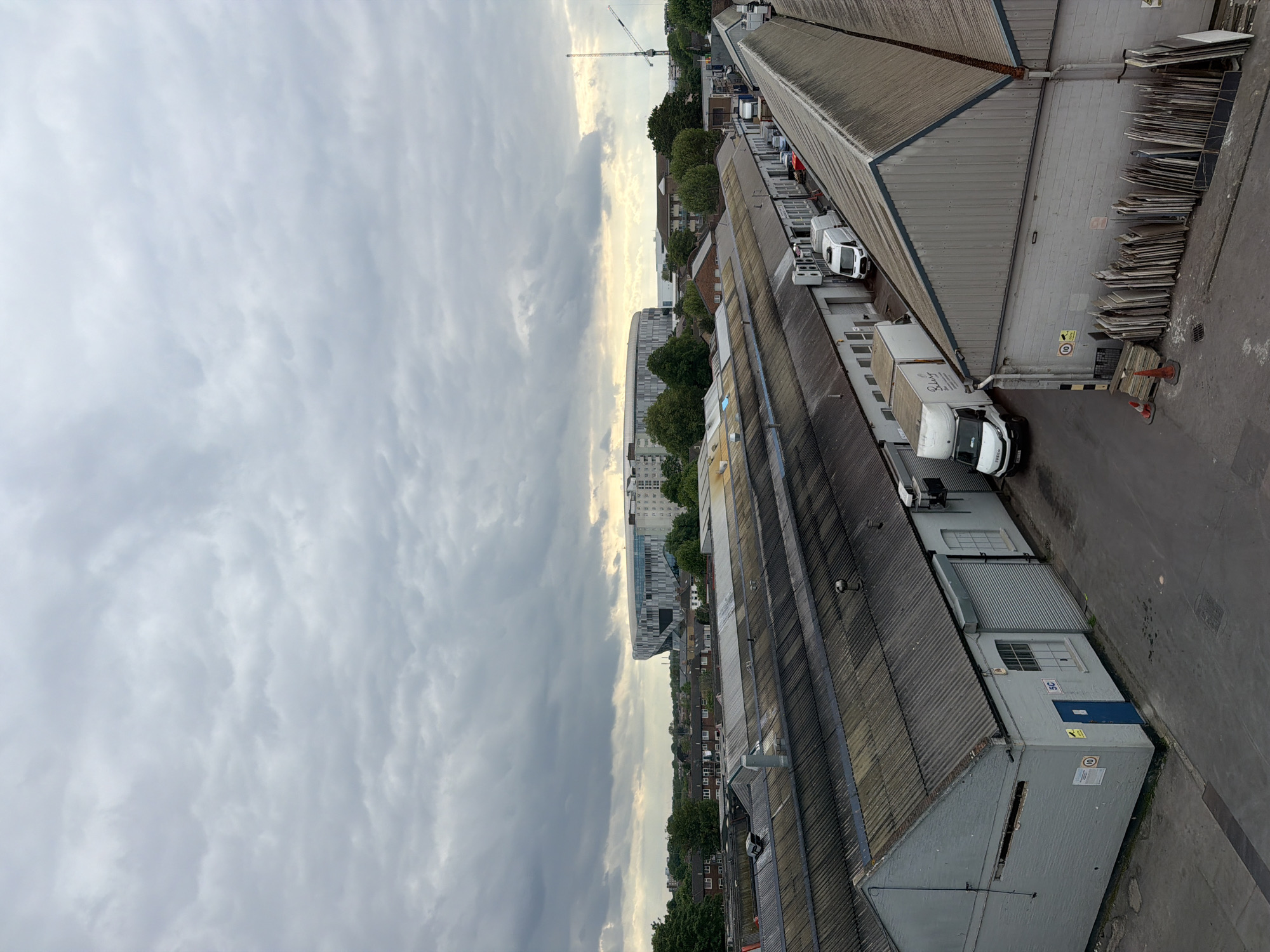 The image shows an aerial view of an industrial or commercial area in London, United Kingdom. A series of long, grey-roofed buildings dominate the foreground, with delivery trucks parked alongside some of them. Piles of construction materials lean against one building. Beyond these buildings, a skyline of residential buildings, interspersed with trees, recedes into the distance. Tottenham Hotspur Stadium is identifiable in the mid-ground. The sky is overcast with thick cloud cover. A construction crane is visible in the background.