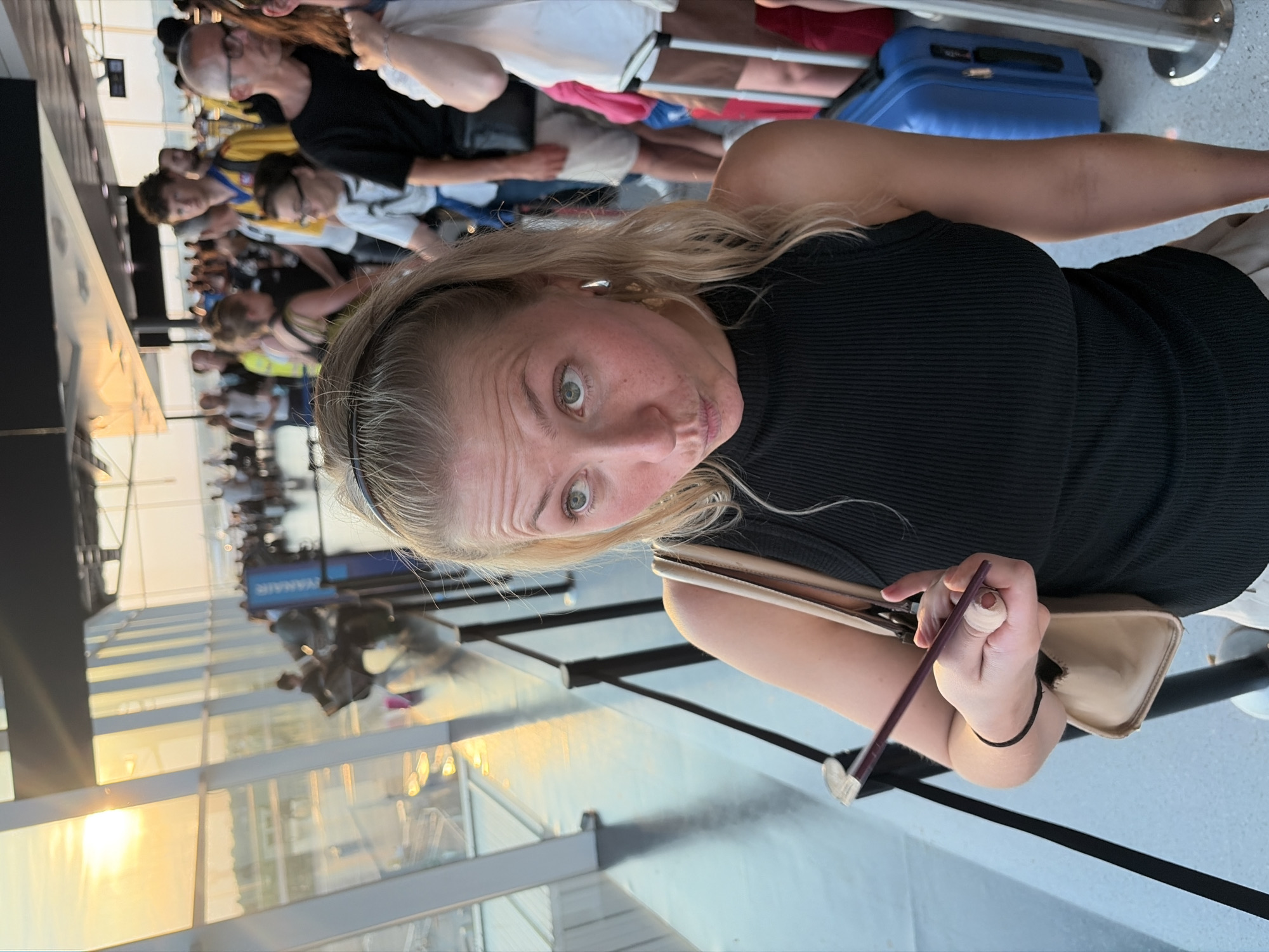 The image captures a group of people in what appears to be an airport terminal or a similar travel hub, given the presence of luggage and a long, reflective walkway. The perspective is close-up on a woman in the foreground, who is looking directly at the camera with a playful, slightly puckered expression. She has blonde hair and is wearing a black top. Her right hand is visible, holding a thin, dark object, possibly a pen or a stylus.
Behind her, other individuals are visible, some in casual attire, suggesting they are either waiting or in transit. A blue suitcase is prominently displayed in the mid-ground. The setting features large glass windows reflecting the sky and the interior of the building, creating a bright and airy atmosphere. The sunlight streaming through the windows suggests it is either morning or late afternoon. The overall mood conveyed by the woman's expression and the casual setting is one of relaxed travel. There is a sign visible in the distance with the text "RYANAIR," which is a low-cost airline, further indicating an airport environment. The setting's polished floors and modern architecture are also typical of airport terminals.