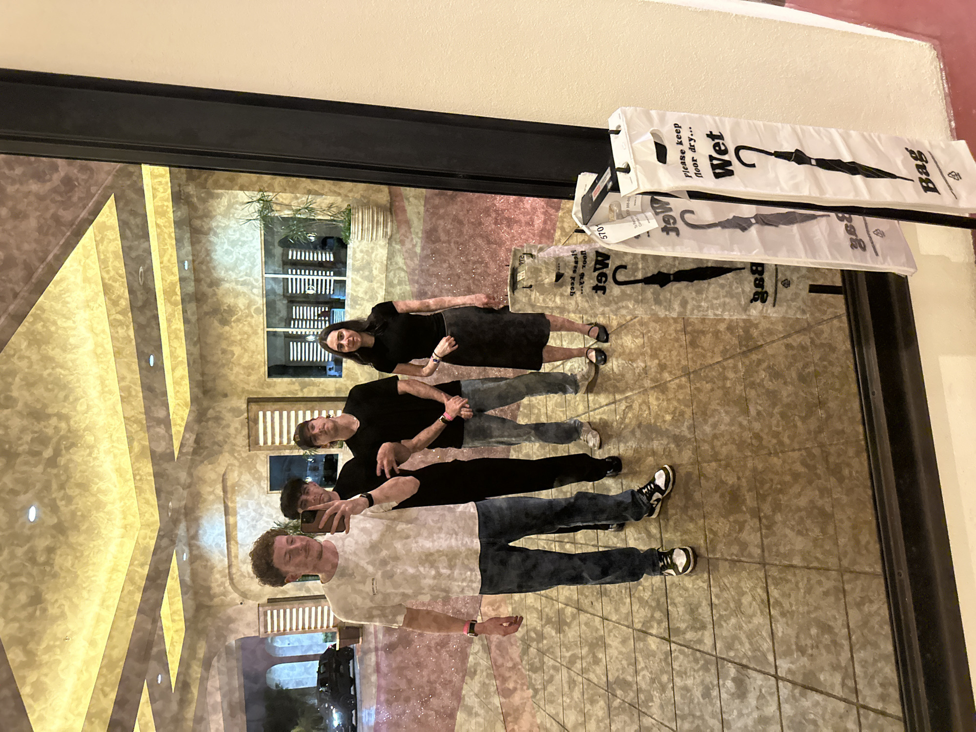 The photograph shows a group of four people standing near a mirrored wall. The mirror reflects their images, creating a doubled visual effect. Starting from the front, a man in a light-colored patterned t-shirt and blue jeans is holding a mobile phone, presumably taking a selfie. Behind him is a man wearing a black t-shirt and blue jeans. Further back is another man in a black t-shirt and blue jeans, and lastly, a woman in a black outfit.
Above them on the wall are bags labeled "Wet Bag." The reflections in the mirror show interior architectural details, including tiled floors, patterned walls with gold-colored vertical accents, and a doorway leading to an area with greenery and outdoor lighting.
Based on visual cues such as the interior design and style, it is possible this scene is inside of the Seminole Hard Rock Hotel & Casino in Hollywood, Florida, within the Seminole Reservation in the United States.