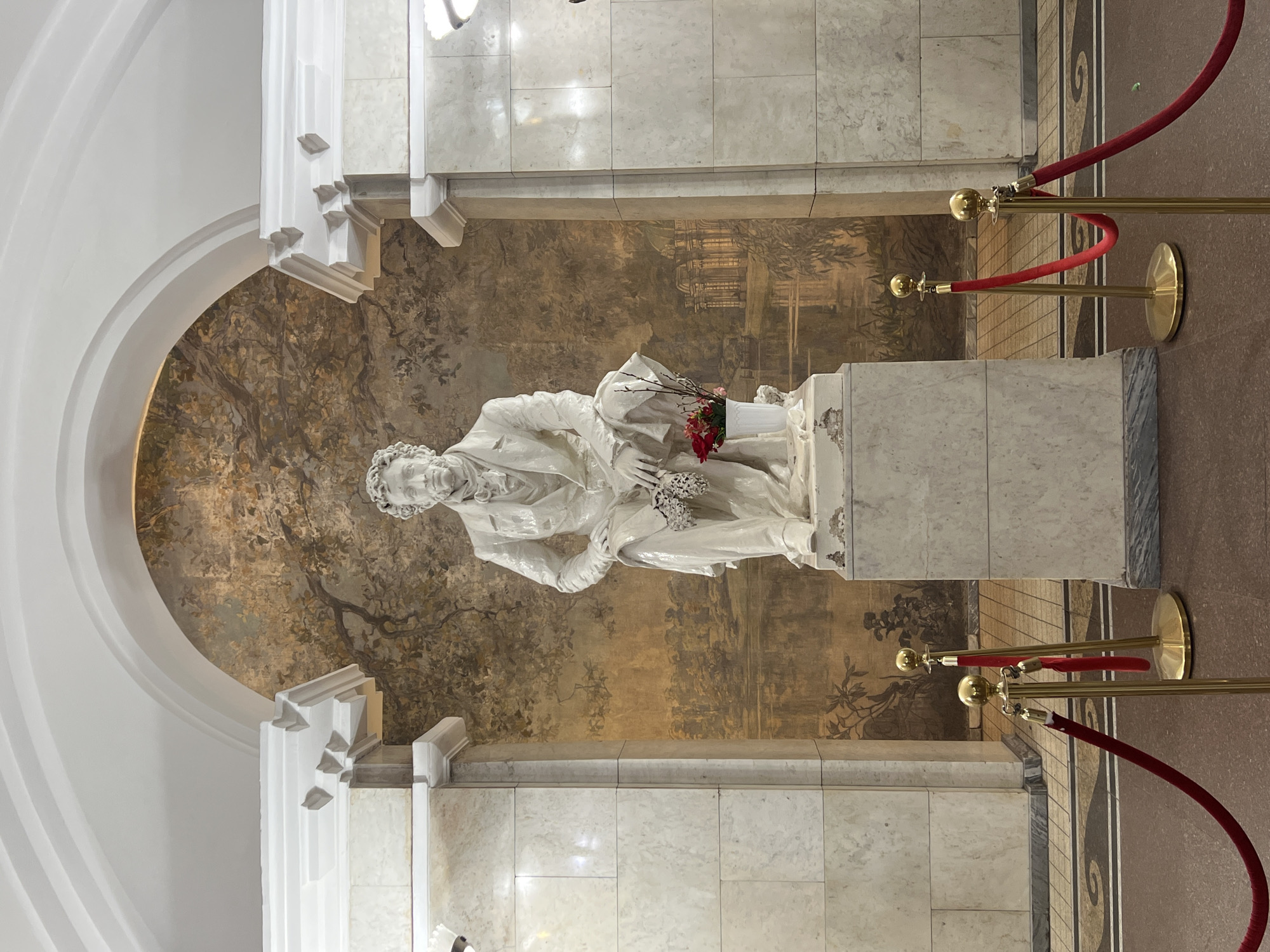 A full shot captures a statue of Alexander Pushkin in Saint Petersburg, Russia. The statue is positioned in front of a mural with a painted landscape of trees, vegetation, and a structure on the far side of the terrain.
The statue is white and surrounded by marble architecture. There is a white arch above the statue. In front of the statue are two red velvet ropes with golden stanchions. The setting appears to be in a museum or monument.