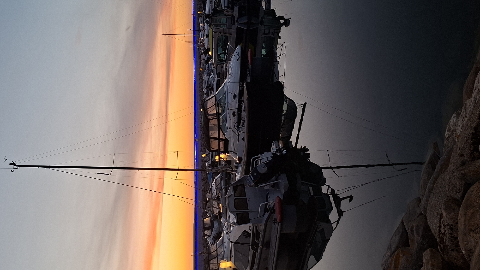 This is a photograph of a marina at dusk, with a sailboat's mast prominently featured in the foreground, casting a reflection on the still water. Several other boats are moored in the background, their silhouettes softened by the fading light and the glowing blue lights lining the harbor. The sky transitions from a warm orange and yellow horizon to a pale blue overhead, with wispy clouds adding texture. Rocks are visible along the bottom edge of the frame, suggesting a breakwater or shoreline. The overall atmosphere is calm and serene, capturing the quiet beauty of a harbor at the end of the day.