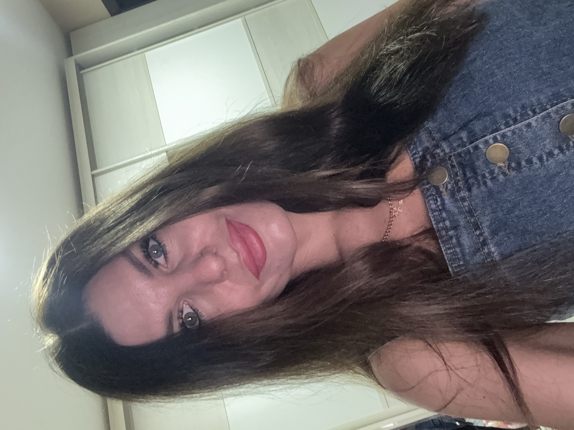 A woman with long, dark brown hair and light blue eyes is pictured in a selfie. She is wearing a denim overall dress and a rose gold necklace. Her lips are painted with a vibrant pink lipstick. The background appears to be a room with a white sliding door wardrobe. The image was taken in Chaikovsky, Russia.