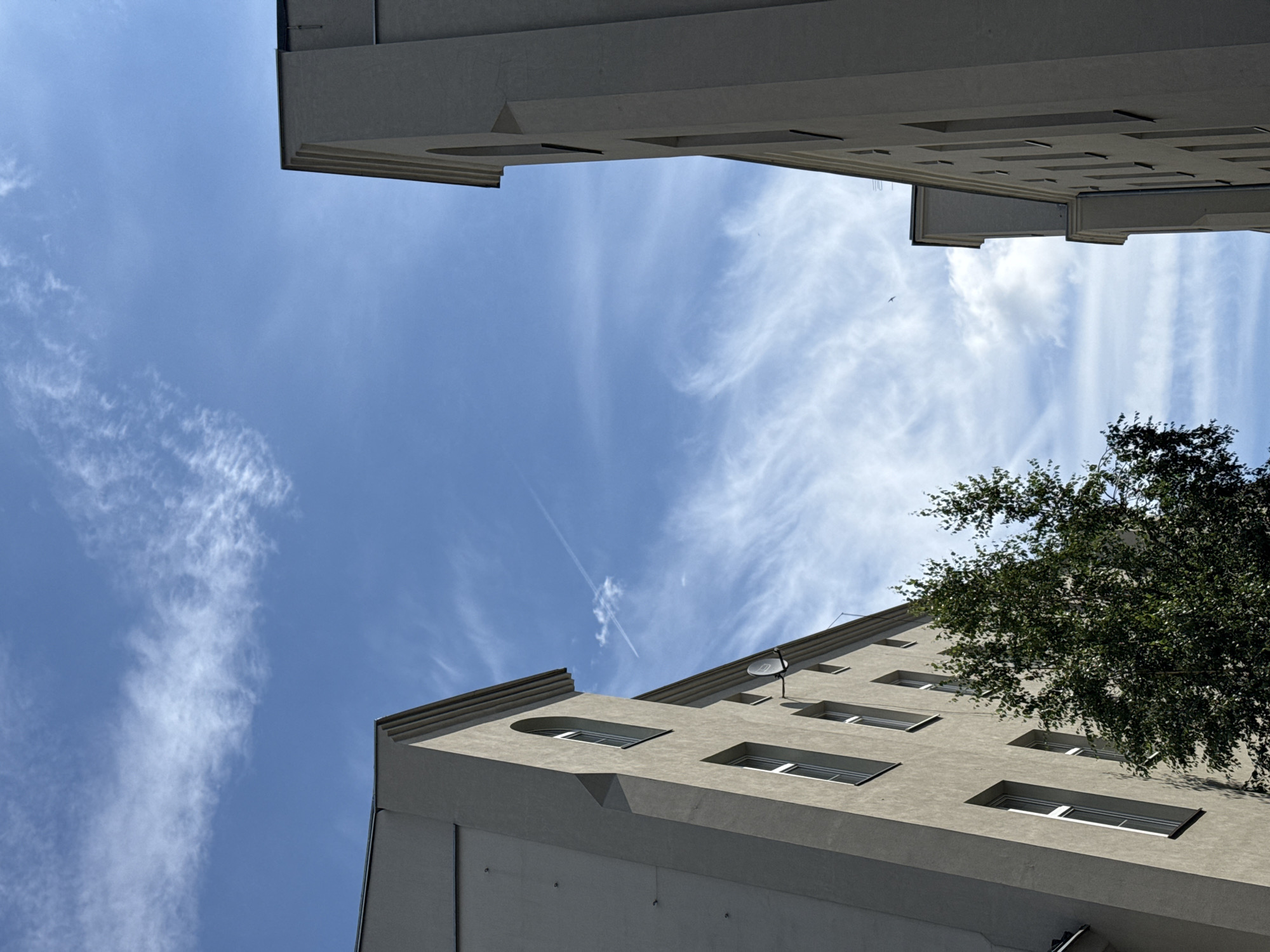 The image displays a low-angle perspective of two modern apartment buildings situated in the city of Łódź, Poland, against a backdrop of a bright blue sky filled with wispy white clouds and a faint contrail from an aircraft. The buildings, constructed from light-colored material, feature rows of uniformly spaced rectangular windows. One building on the left has a distinctive arched window at its peak, and a satellite dish is visible on its facade. A lush green tree partially obstructs the view of the left building. The building on the right is taller and appears to be part of a larger complex. The architecture suggests a contemporary urban setting.